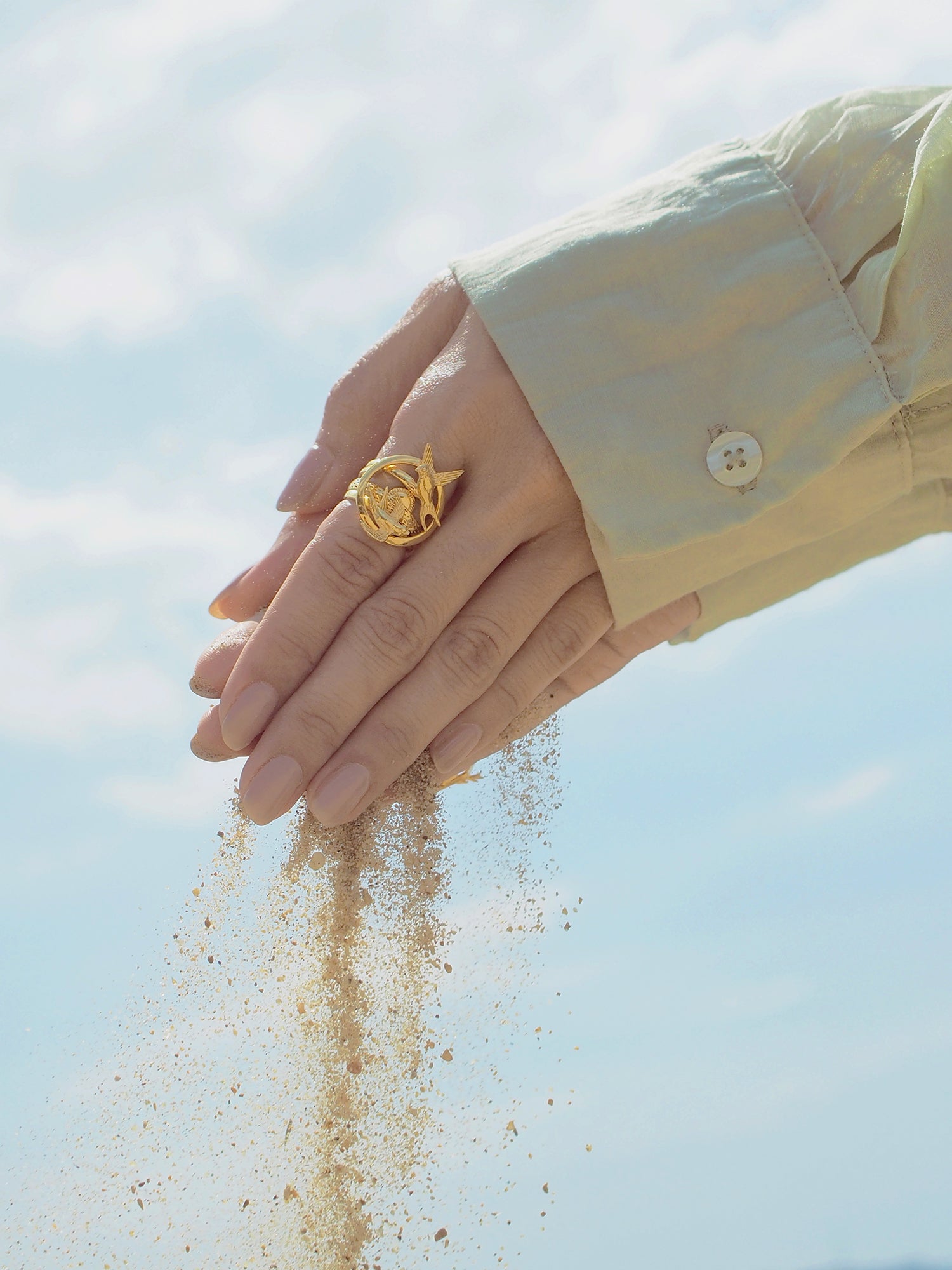 Hand with beautiful stacking rings releasing sand against a blue sky.
