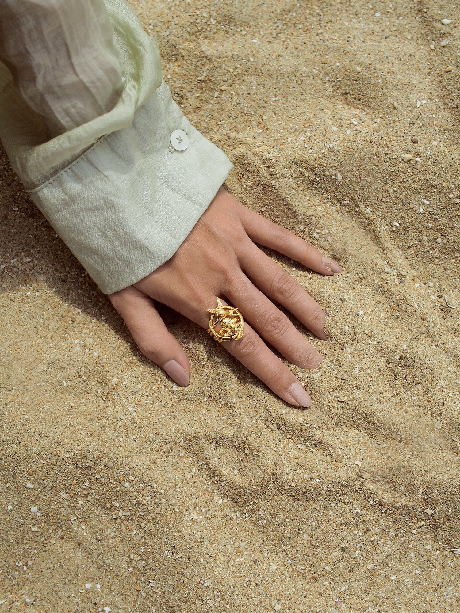 A hand resting on sand, showcasing a gold stacking ring.