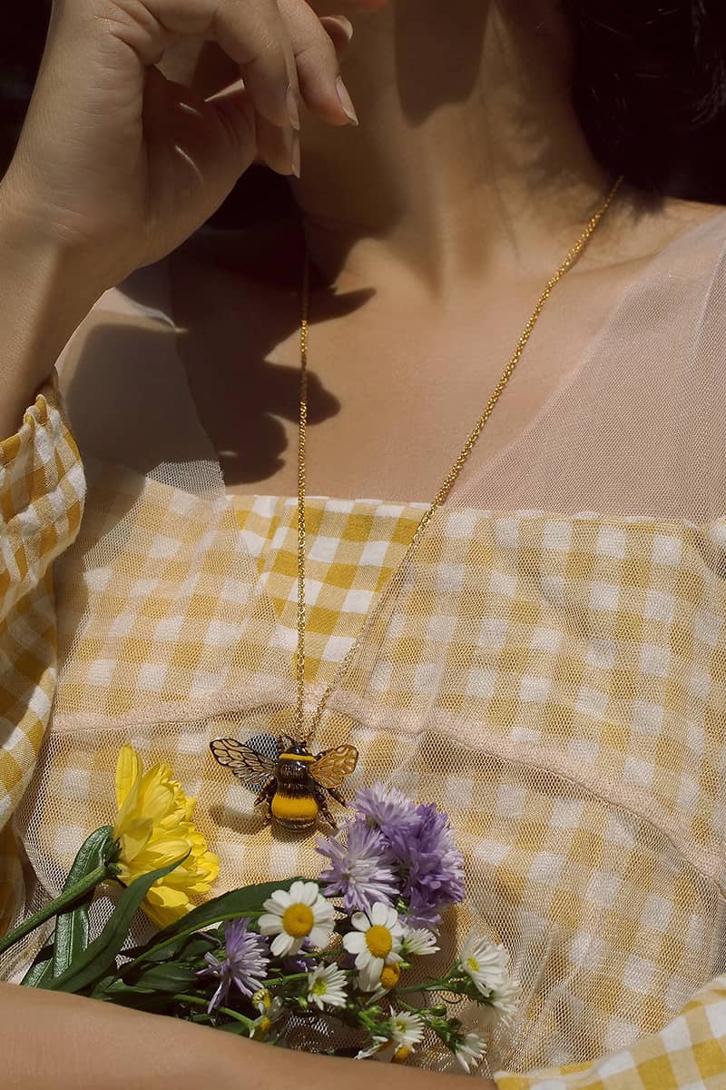 Close-up of a woman wearing a bee pendant necklace with a yellow dress and holding a bouquet of flowers.