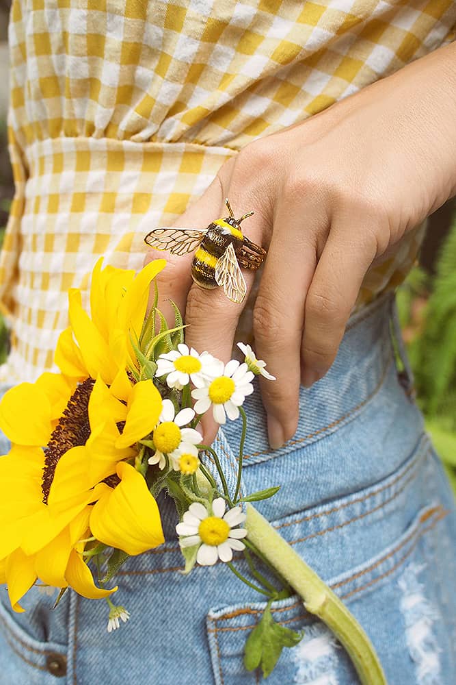 Close-up of a woman's hand adorned with a charming bee, ring surrounded by sunflowers and daisies.