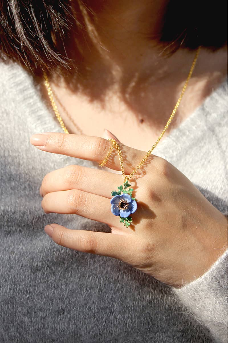 Nature-inspired necklace featuring a purple flower and green leaves pendant on a golden chain, held by a model's hand.