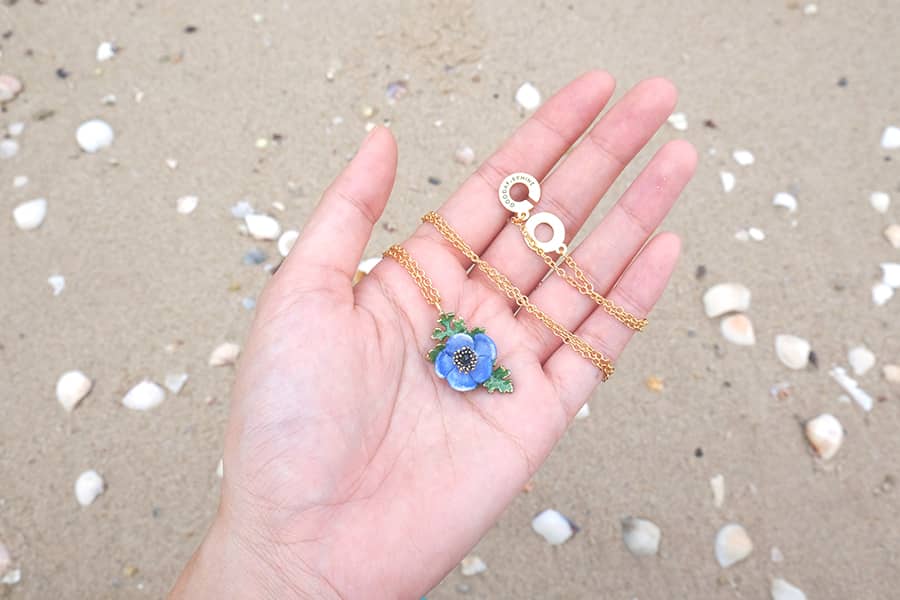 Close-up of a hand holding necklace with a purple flower and green leaves on sandy beach.