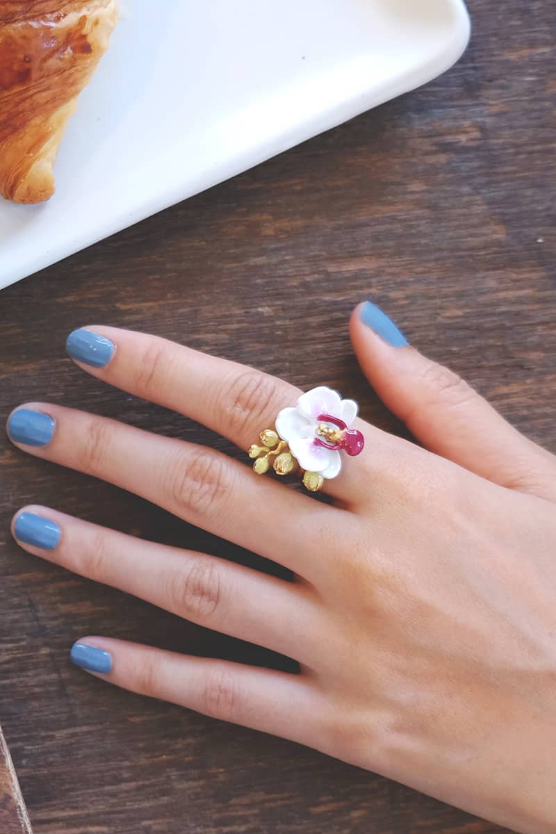 A hand with blue nail adorned with a floral ring featuring a white phalaen design