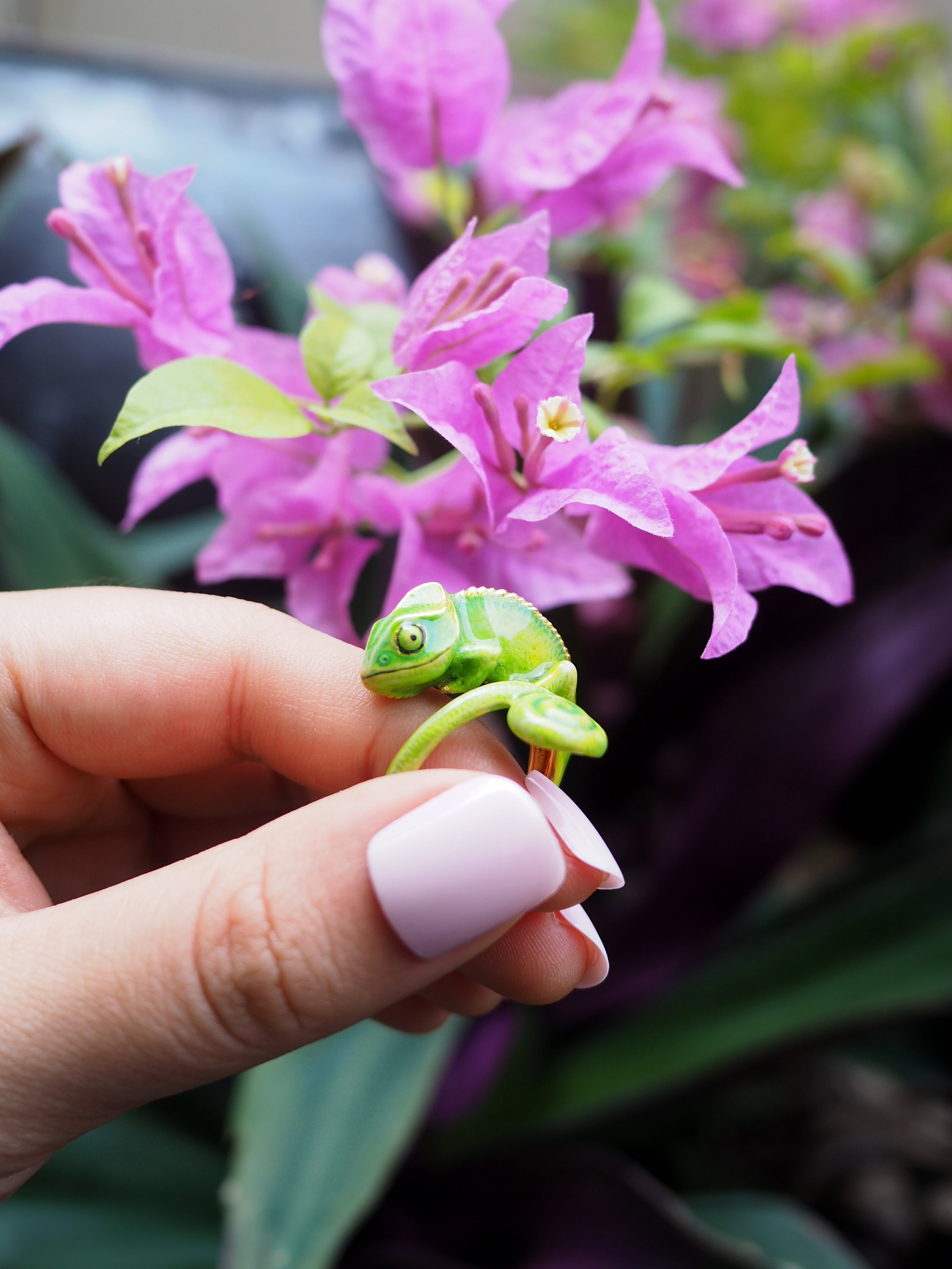 Close-up of a green chameleon ring held in a hand with pink flowers in the background.