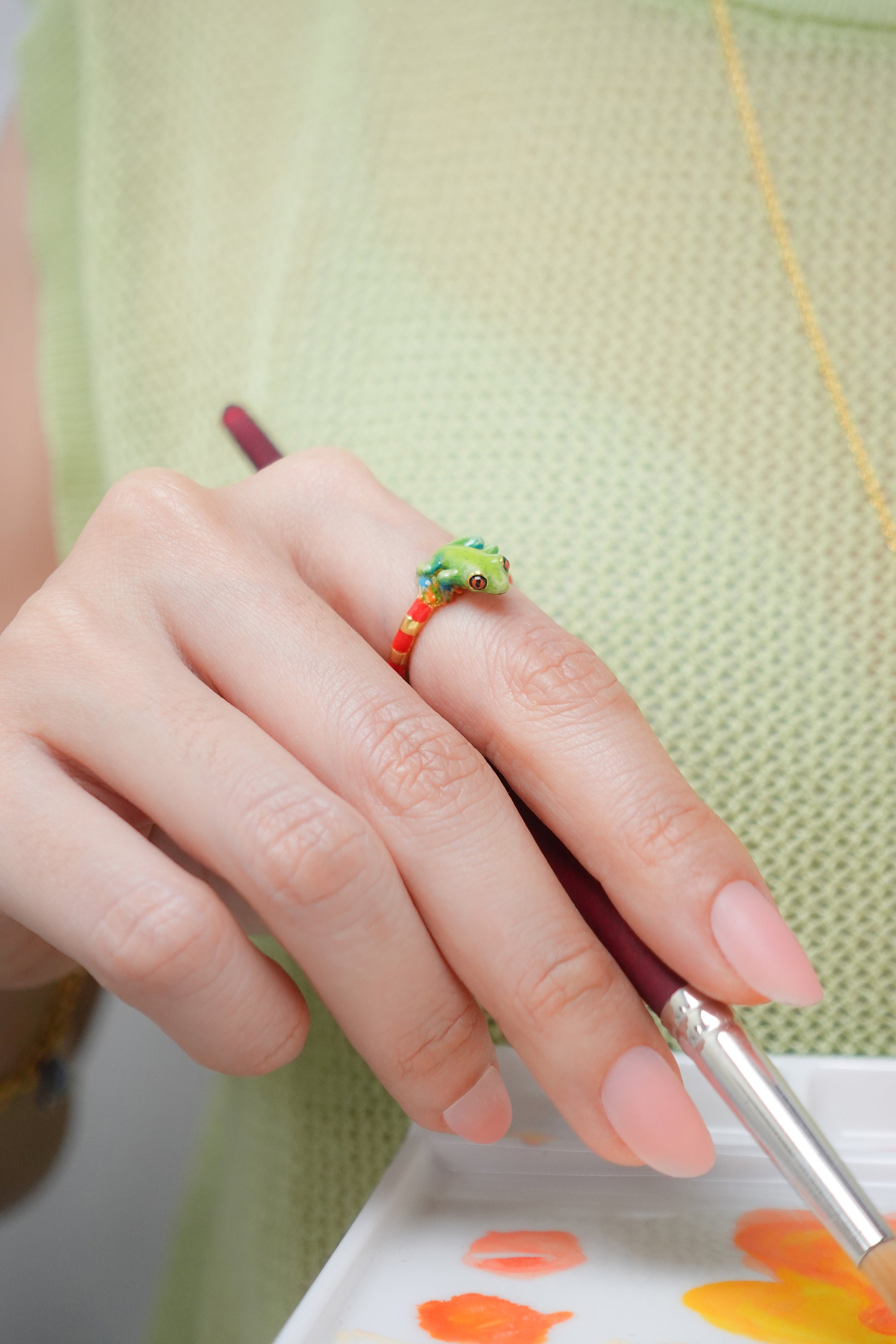 A close-up of a hand wearing a colorful frog ring while holding a paintbrush.
