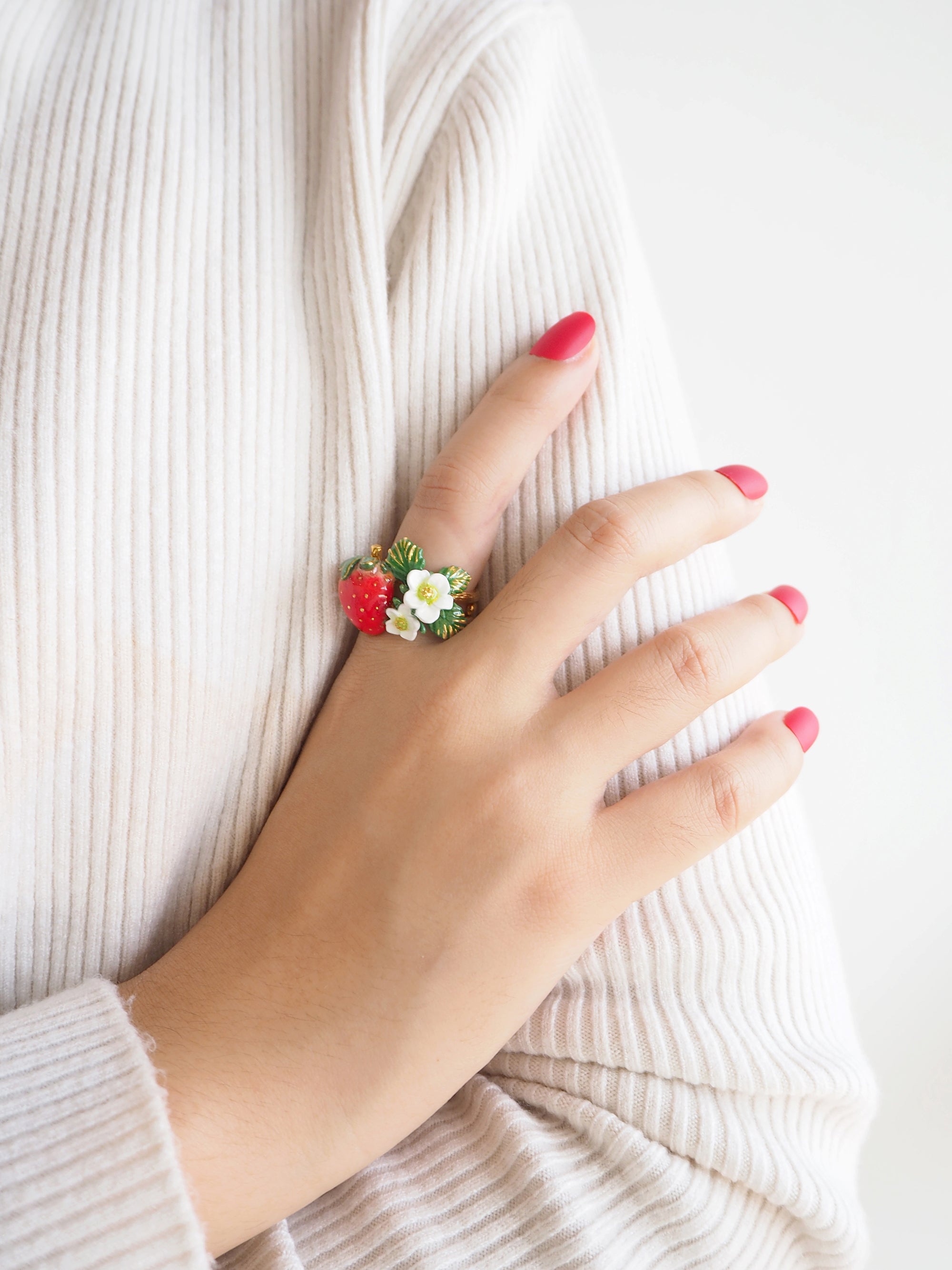 Close-up of a hand with red nails wearing a beige sweater and a beautiful floral strawberry ring