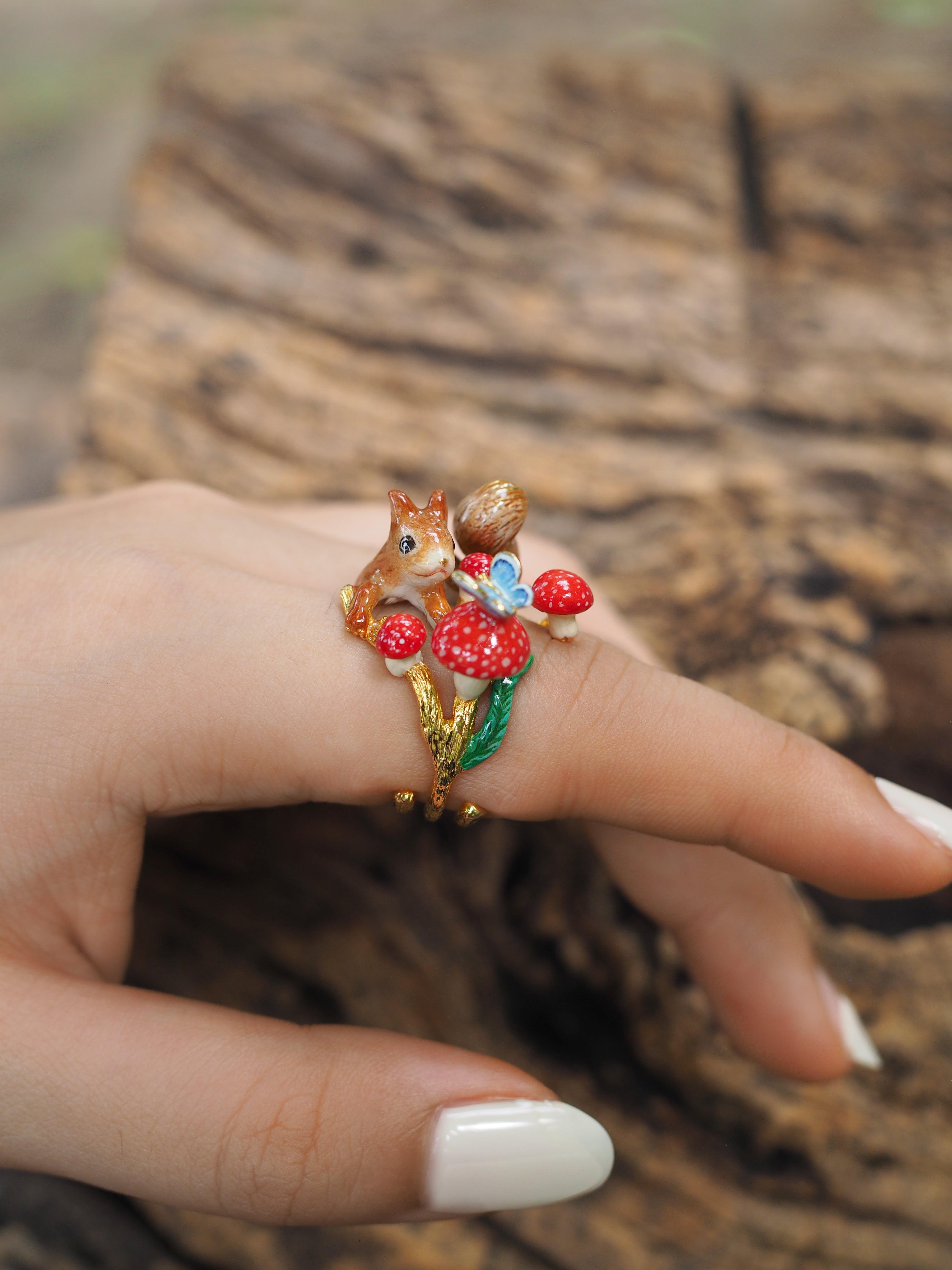 Close-up of a hand adorned with a gold ring featuring a squirrel, mushrooms, and a blue butterfly design against a wooden background.