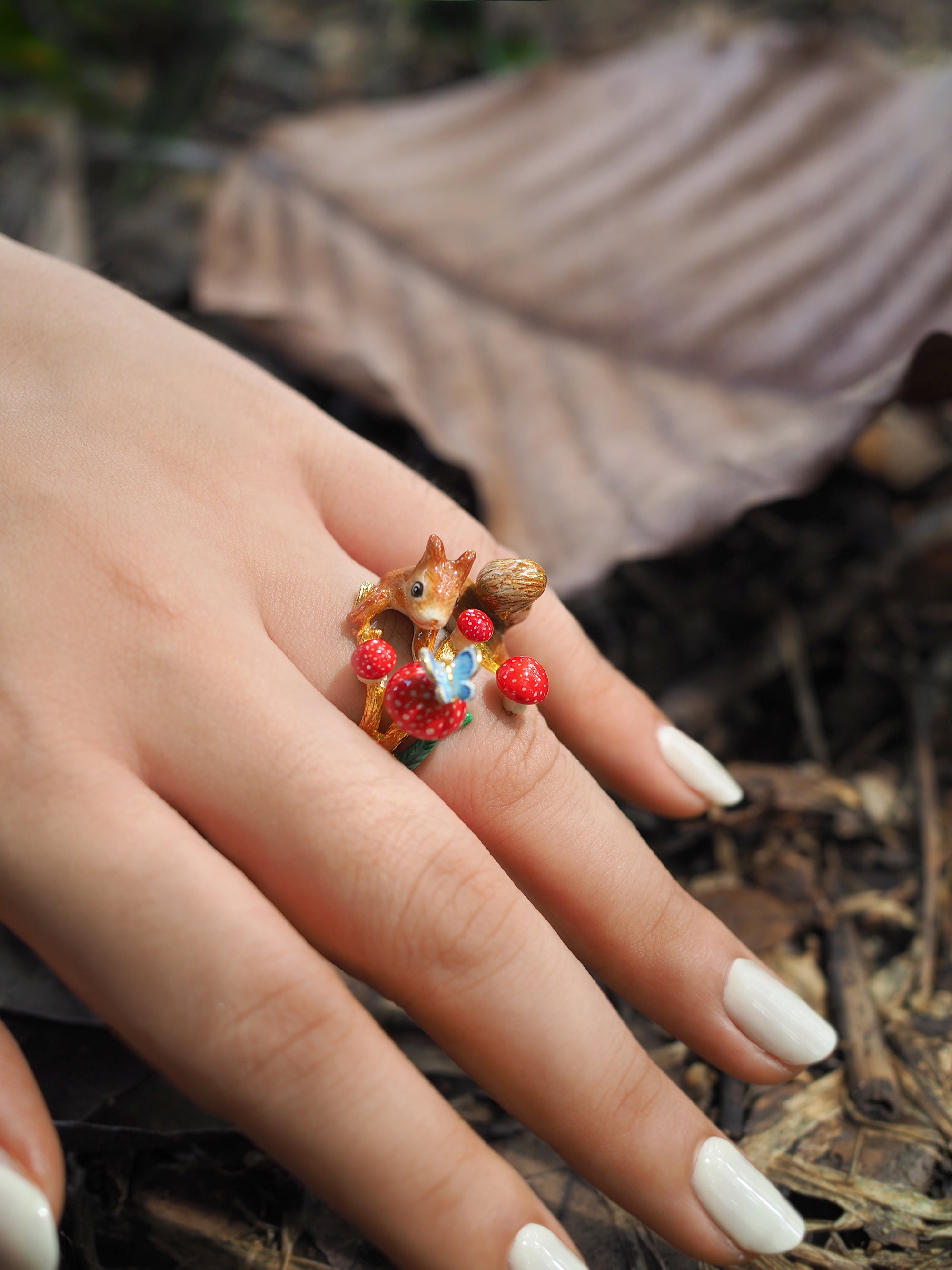 Close-up of a hand wearing a gold ring featuring a squirrel, mushrooms, and a blue butterfly design surrounded by dried leaves.