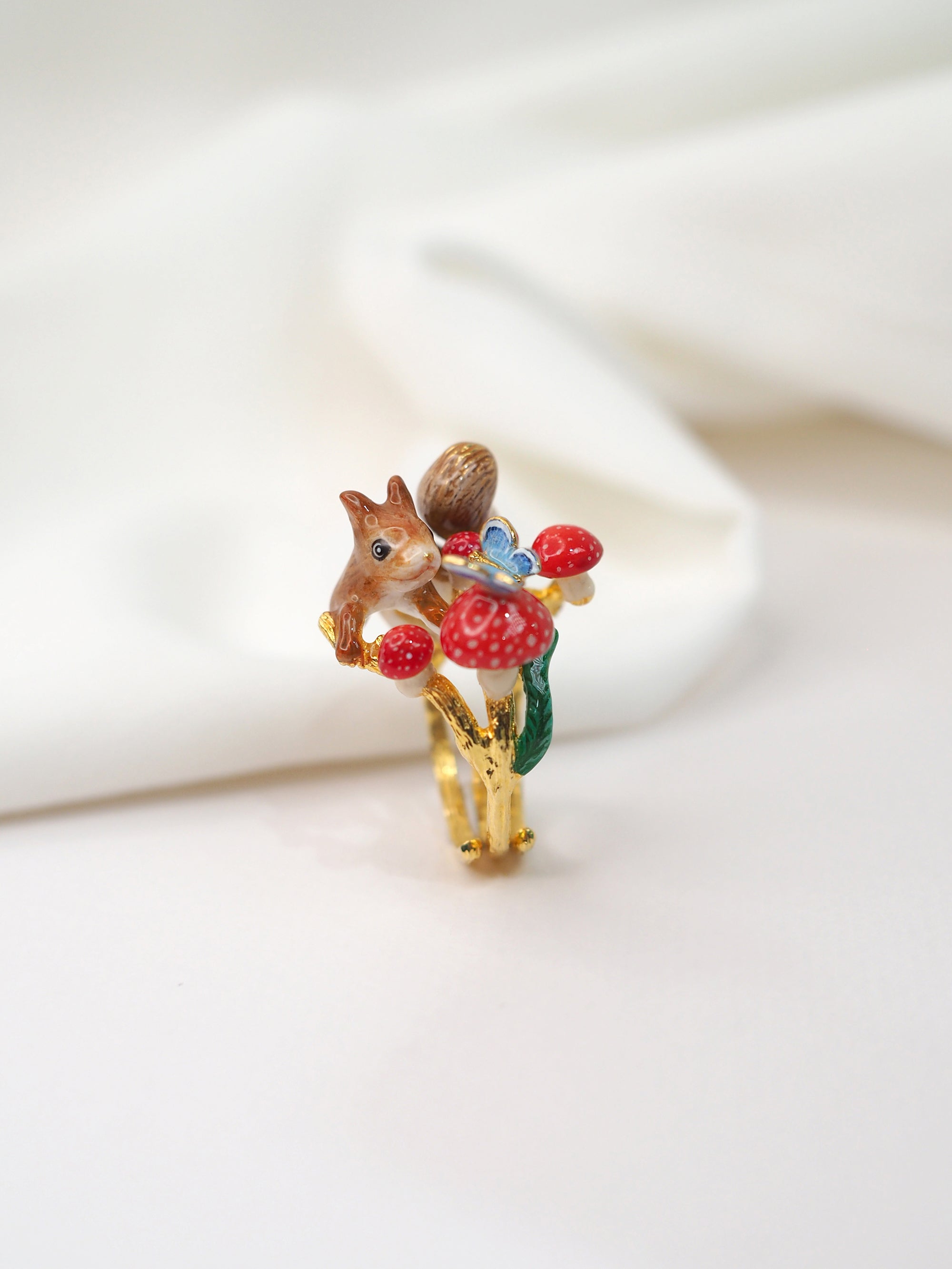 Charming gold ring featuring a squirrel, red mushrooms, and a blue butterfly design placed on a white fabric.