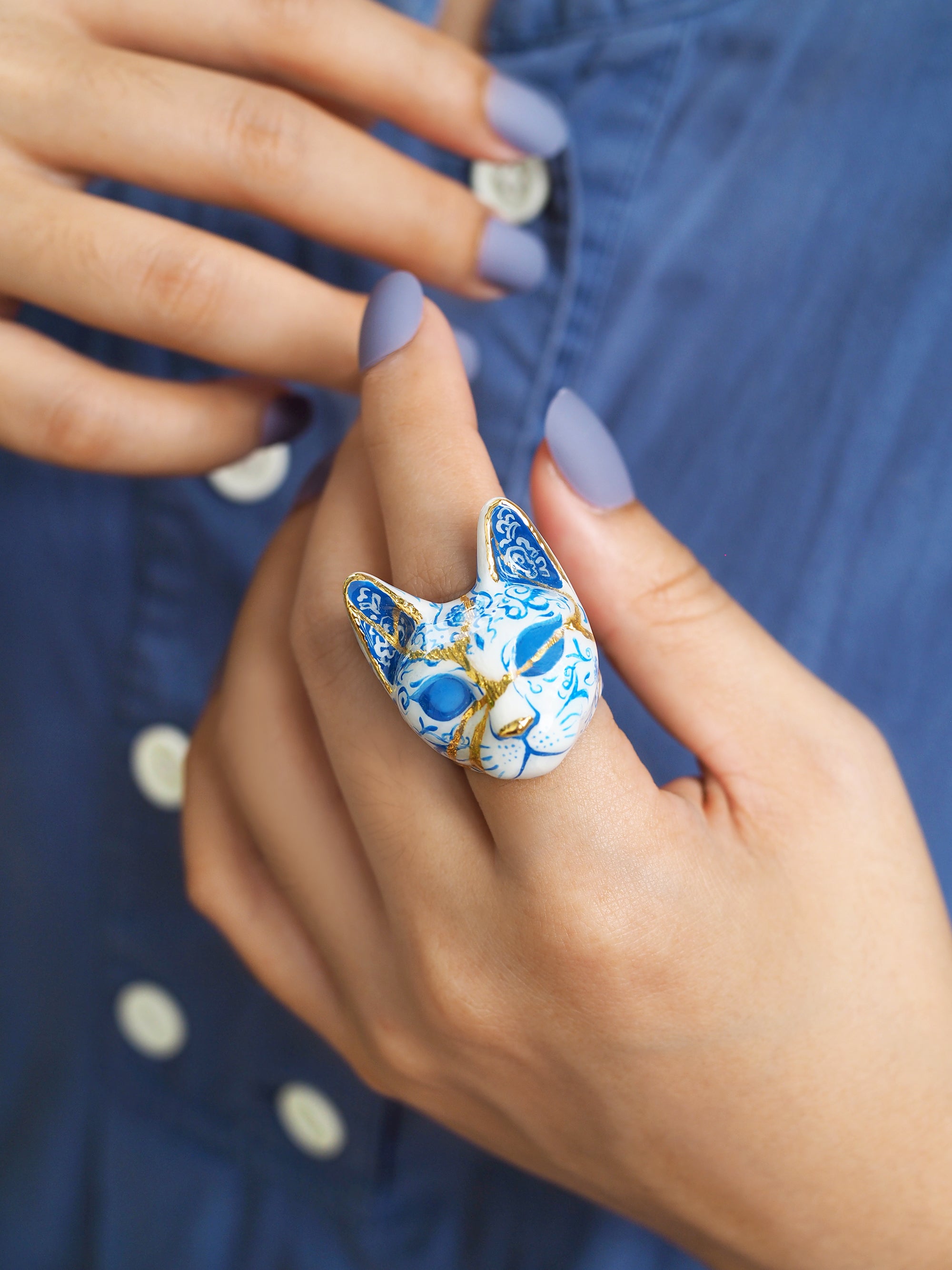 Close up of a hand with lavender nails showcases a blue and white cat ring, detailed with porcelain patterns.