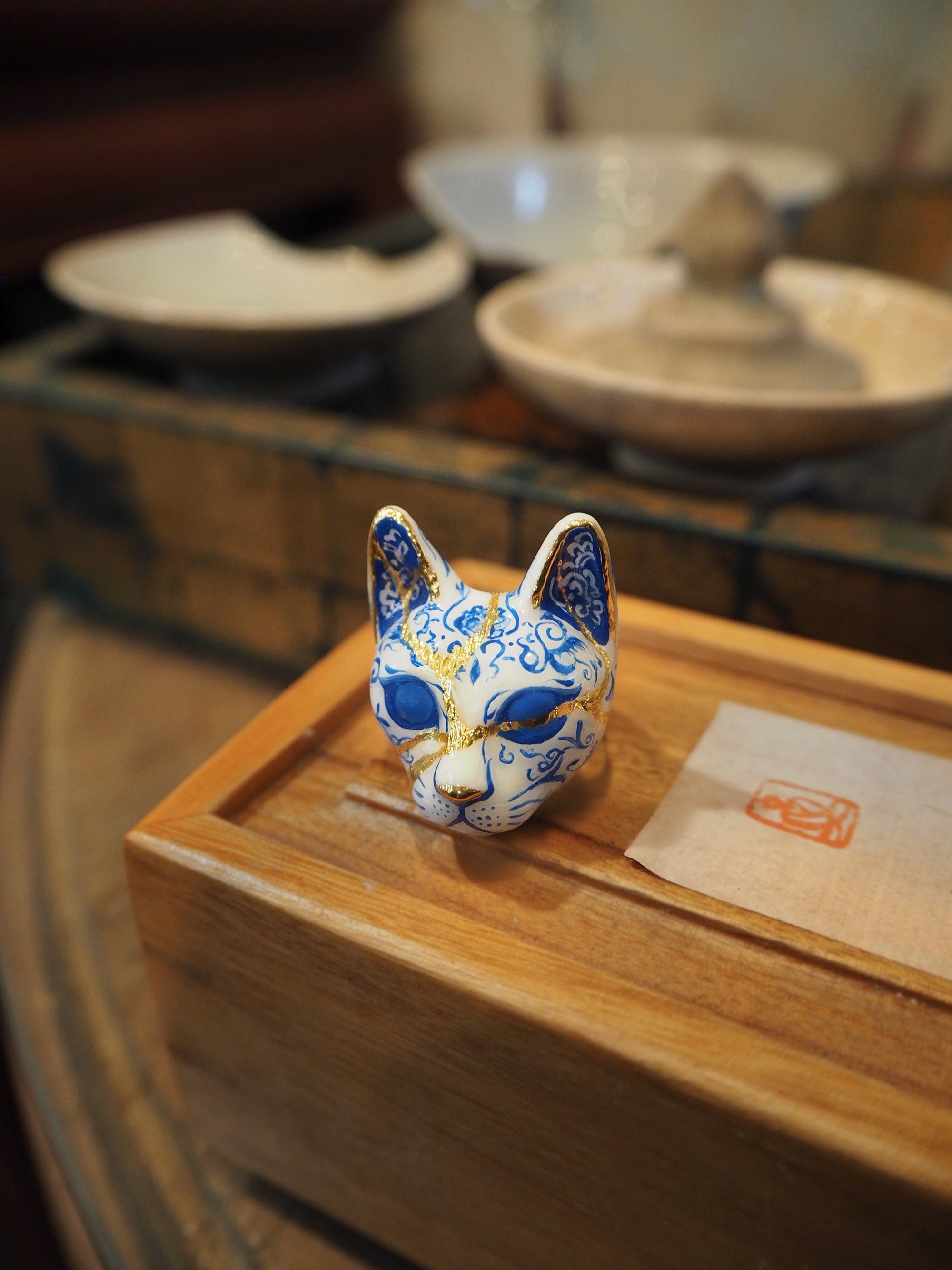 Hand-paint cat's face ring featuring detailed porcelain patterns, painted in blue and white pattern with gold accents, place on a wooden box.