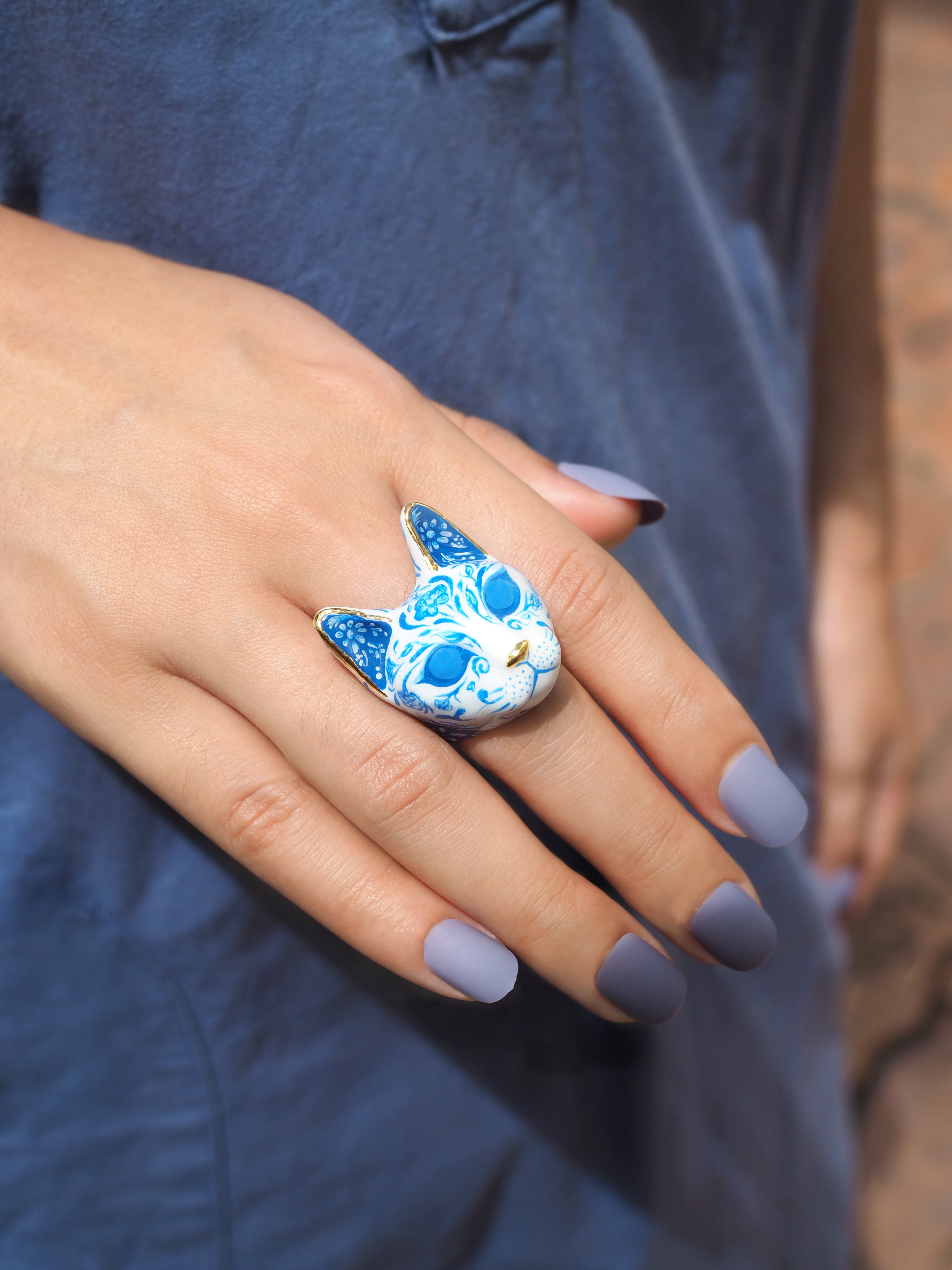 A close-up of a hand with a blue and white cat-shaped ring and light purple nails, wearing a casual blue shirt in natural light.