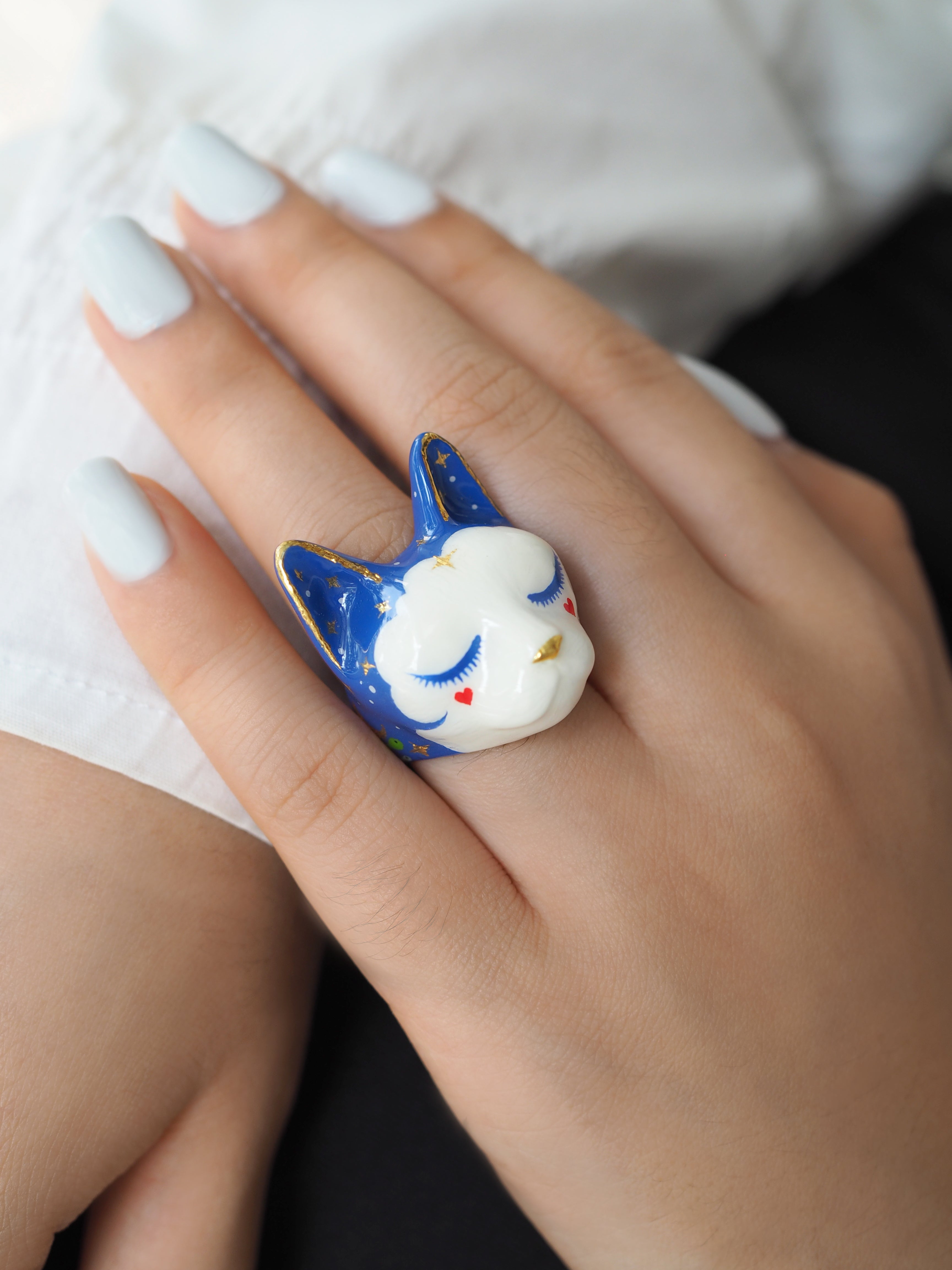 Close-up of a whimsical cat ring featuring a blue design and cute facial details worn on a woman's hand.