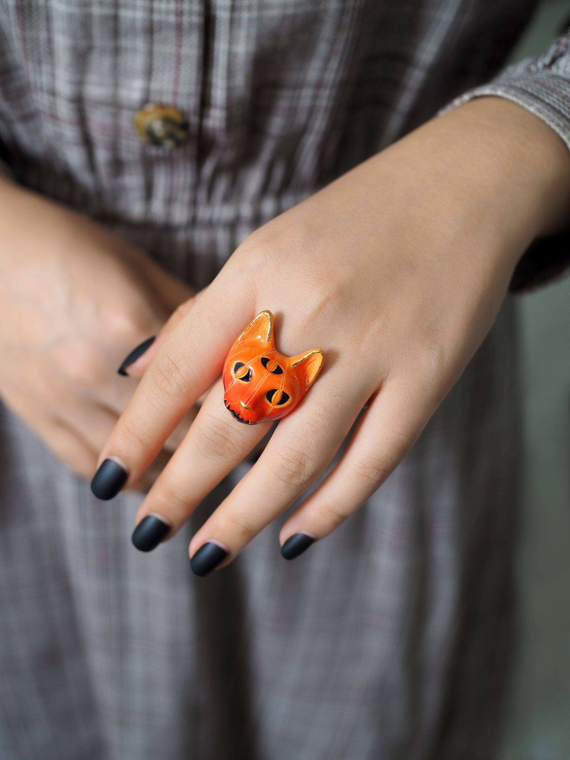Halloween-themed ring shaped like a bright orange cat's face, worn on a hand with dark-painted nails.