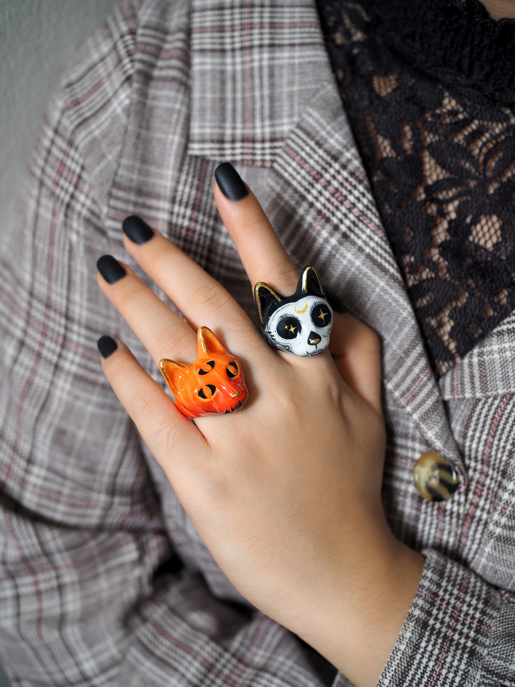 A close-up of a hand with black nail polish, adorned with two Halloween-themed cat rings a black cat with a skull motif and a pumpkin.