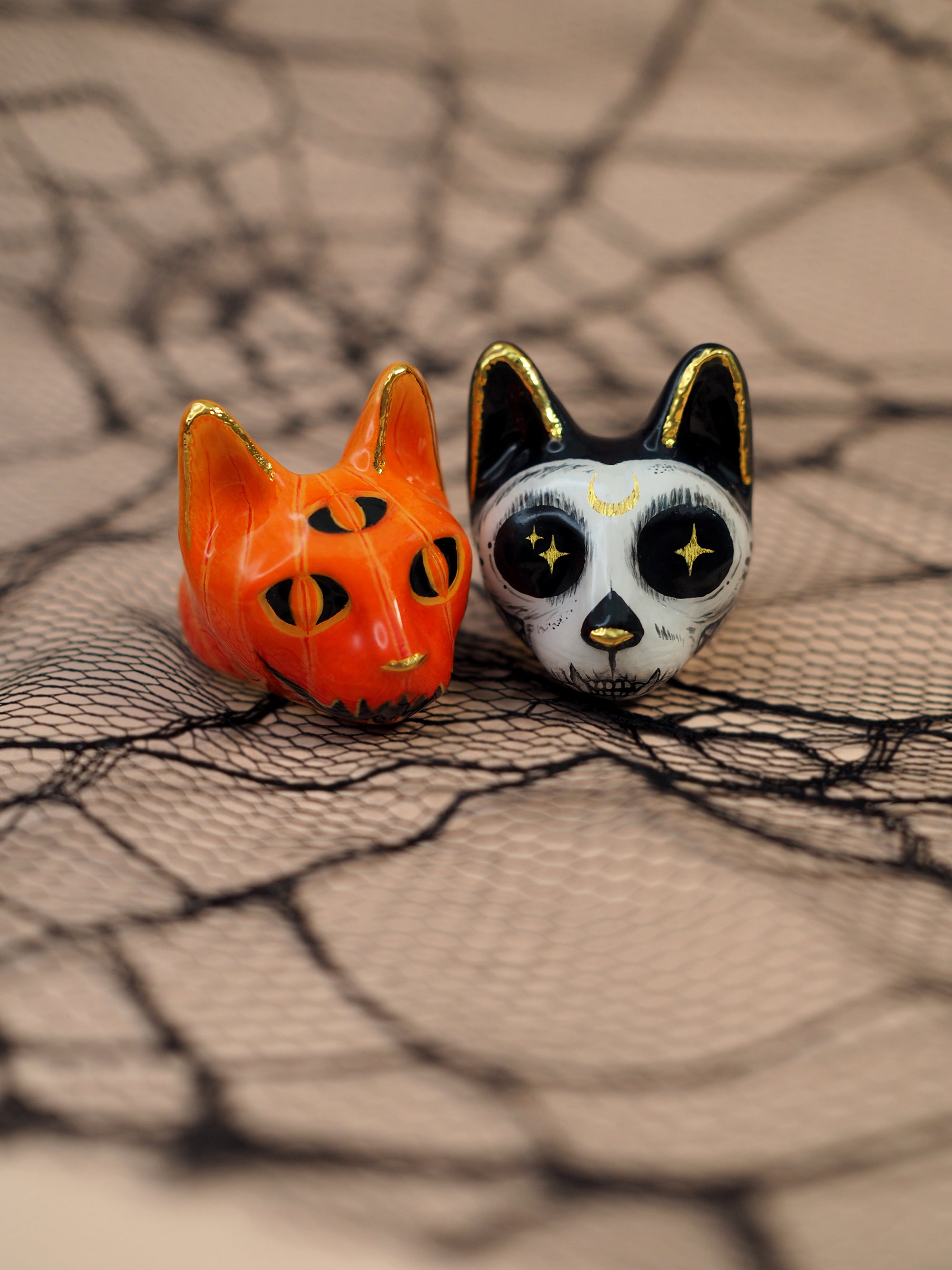 A close-up of two Halloween-themed cat rings a black cat with a skull motif and a pumpkin et on decorative spider web.