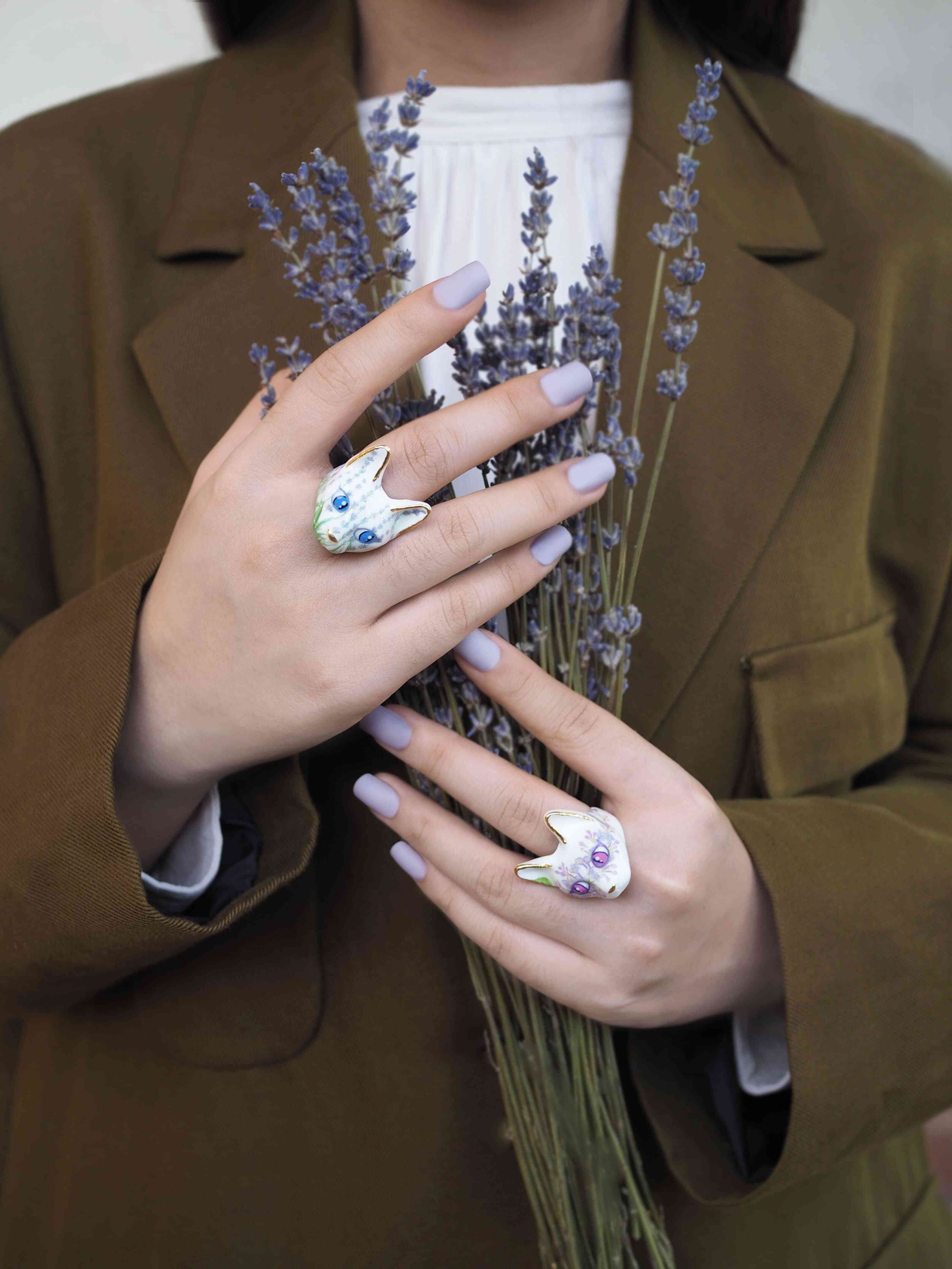 A person holds lavender flower with hands adorned in unique cat rings featuring floral pattern, with soft pastel nails and an olive jacket.