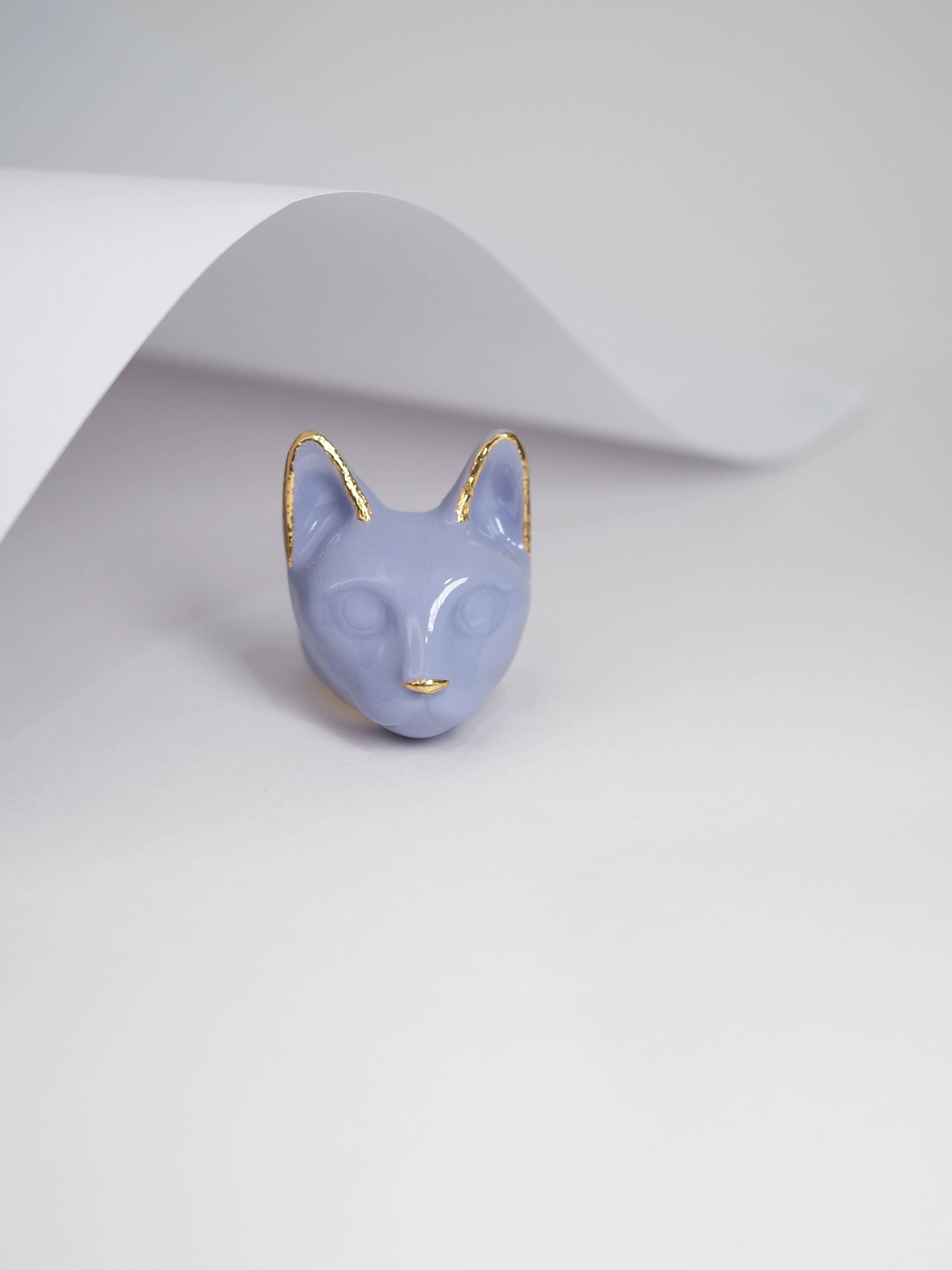 A whimsical ring designed in the shape of a cat’s head in purple placed on a white background, perfect for art jewelry collectors.