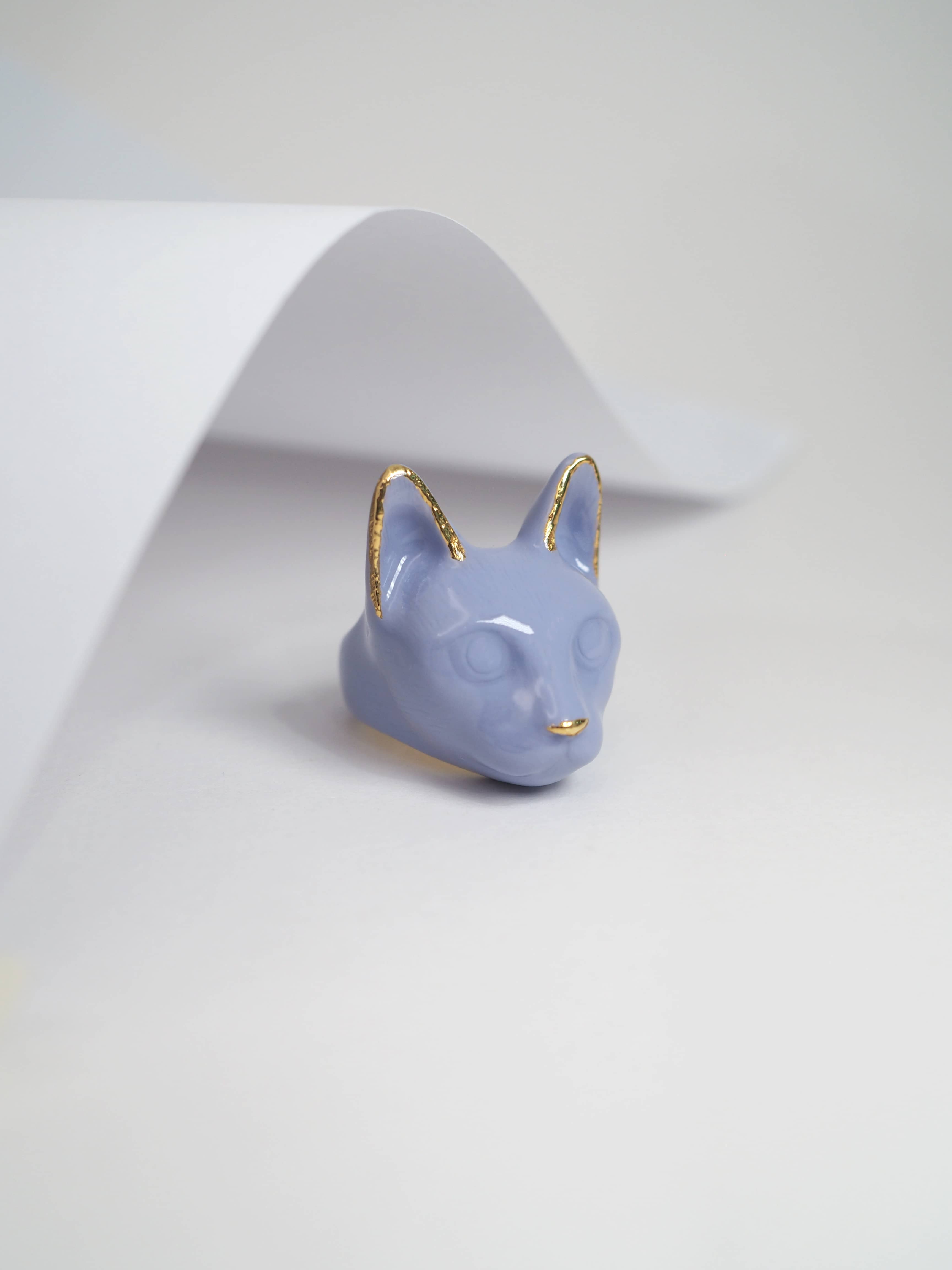 A whimsical ring designed in the shape of a cat’s head in purple placed on a white background.