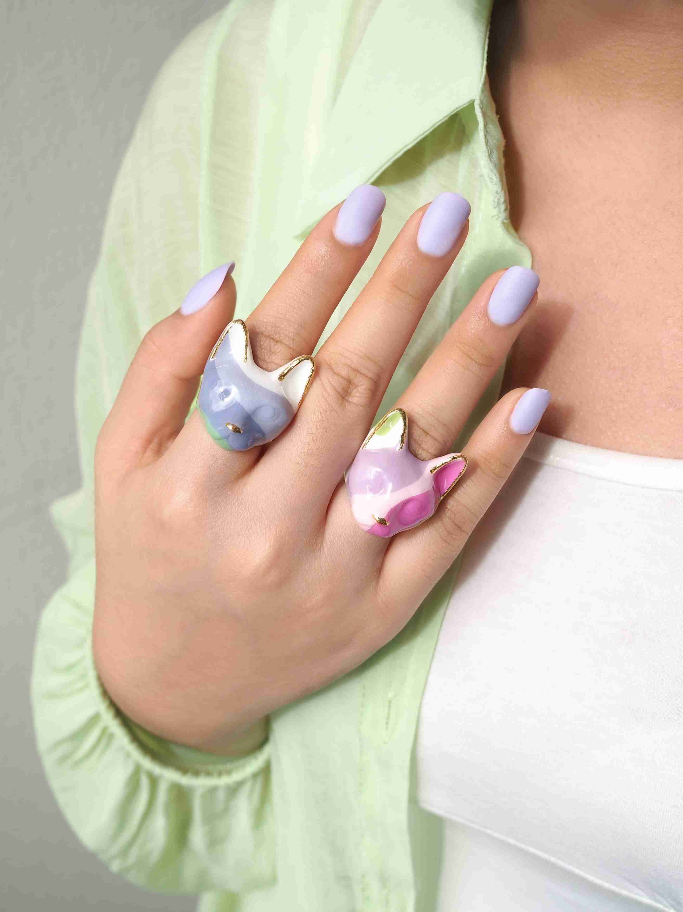 A hand with lavender nails displays two cat rings in pastel color, purple, pink and green.
