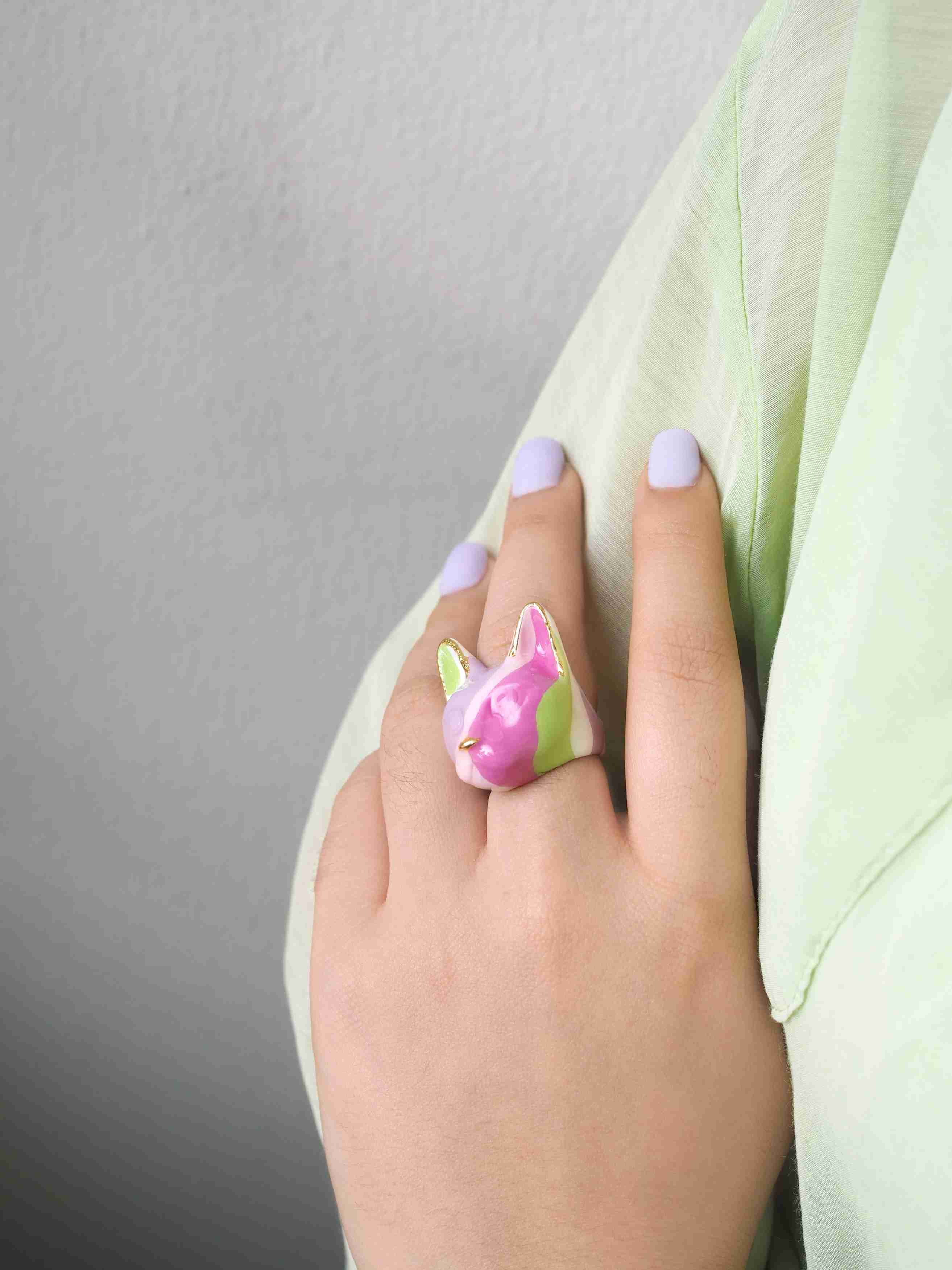 Close up of a hand adorned with whimsical cat ring in purple, pink, green, and white, ideal for summer look.