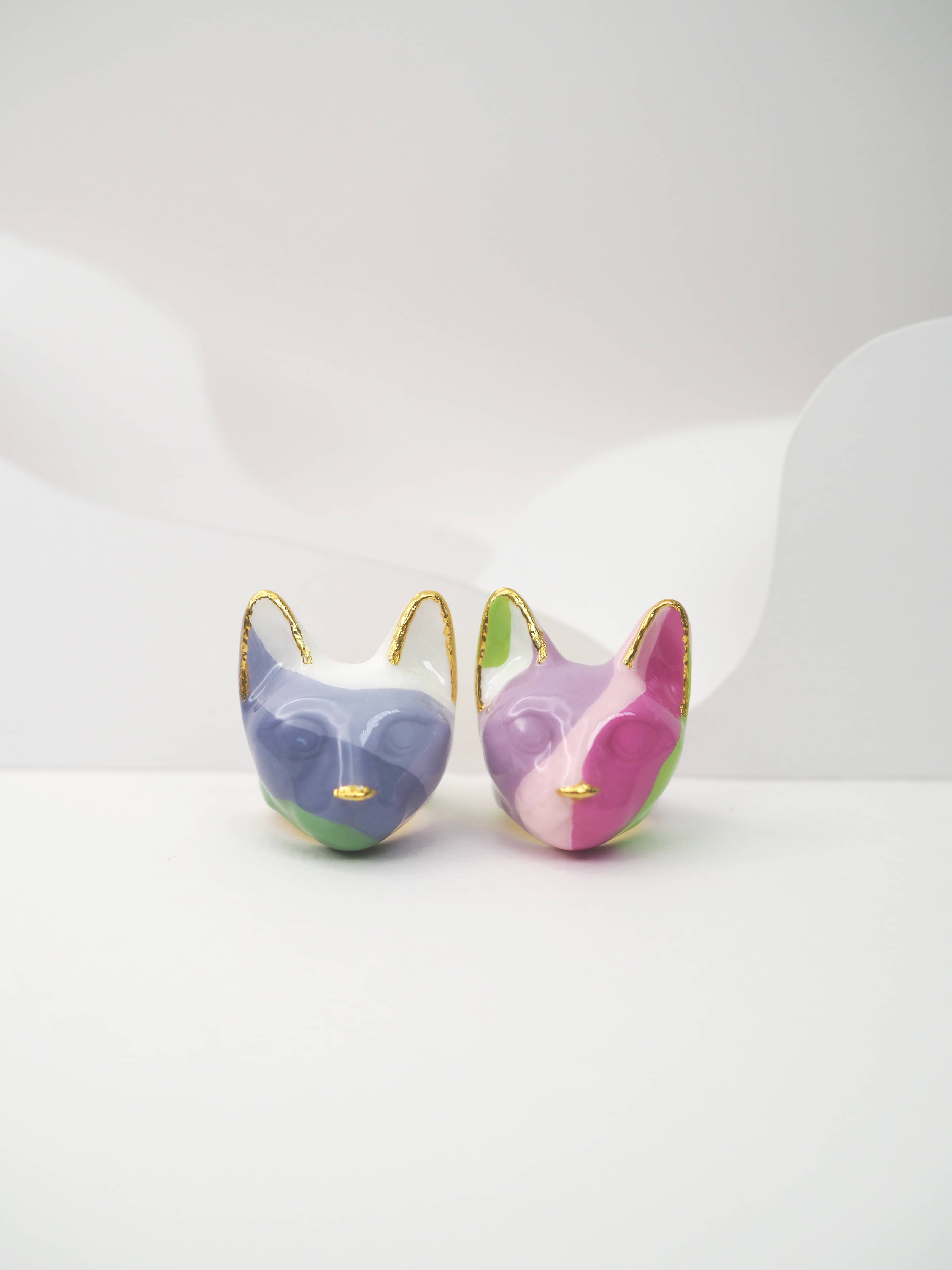Two unique cat ring in pastel colors—one purple, green, and white, the other pink, green, and white—set against a light background.