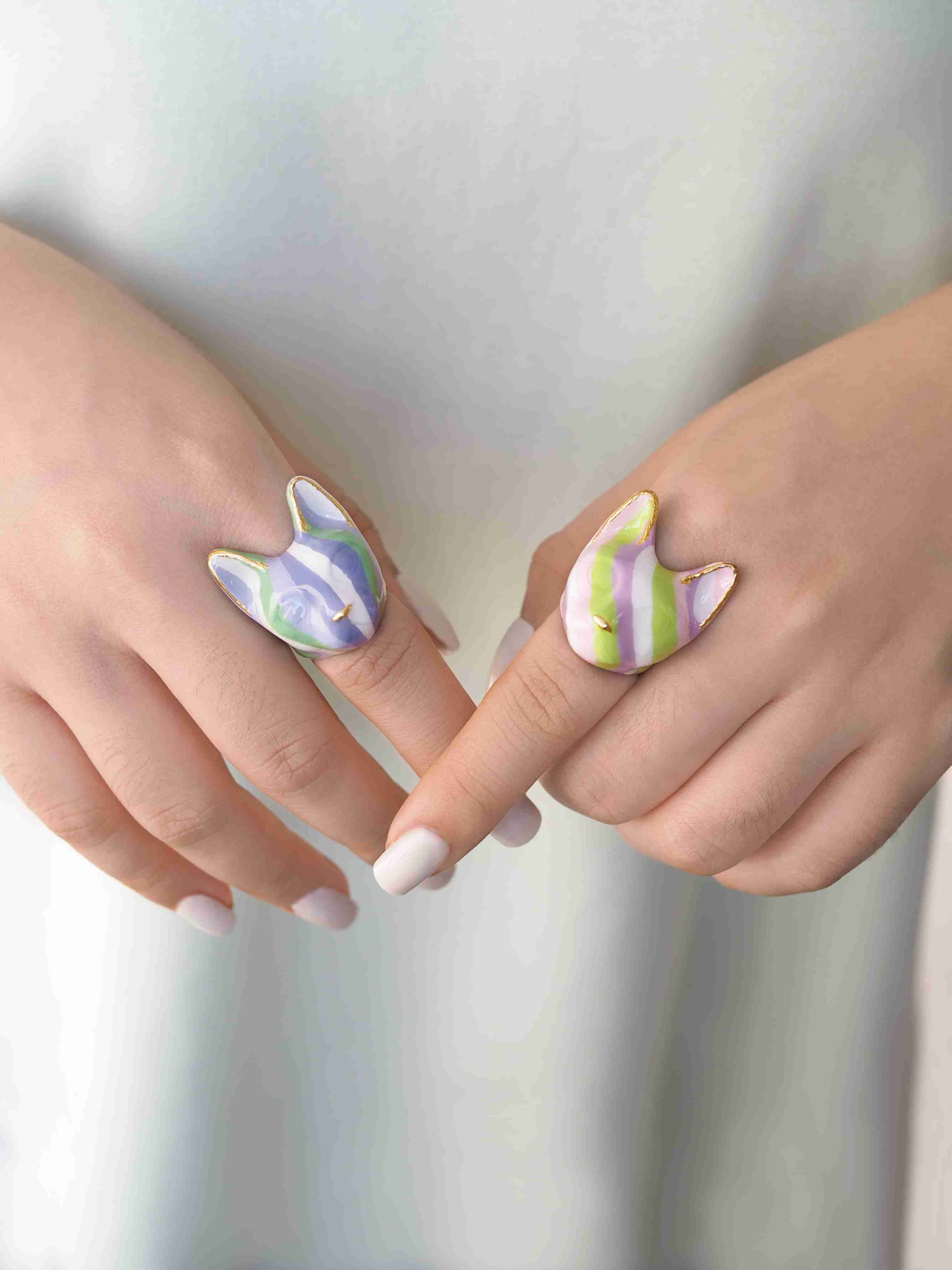 Two hands are shown wearing playful cat-shaped rings with soft purple, green, and white stripes, while the other features green, pink, purple, and white stripes.