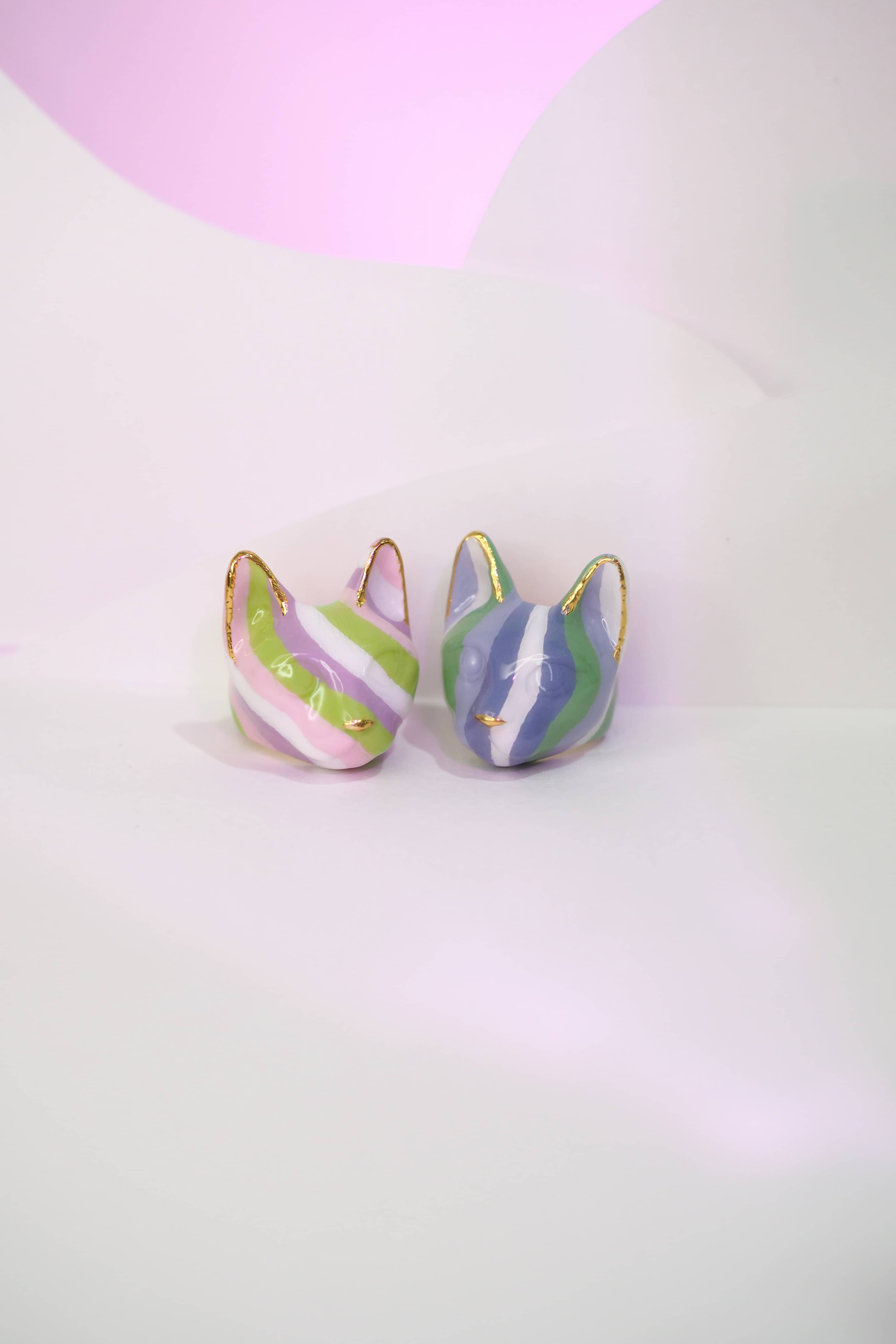 Two cat rings features alternating stripes of purple, green, and white, while the other with lilac, light green and pink set against a soft pastel background.