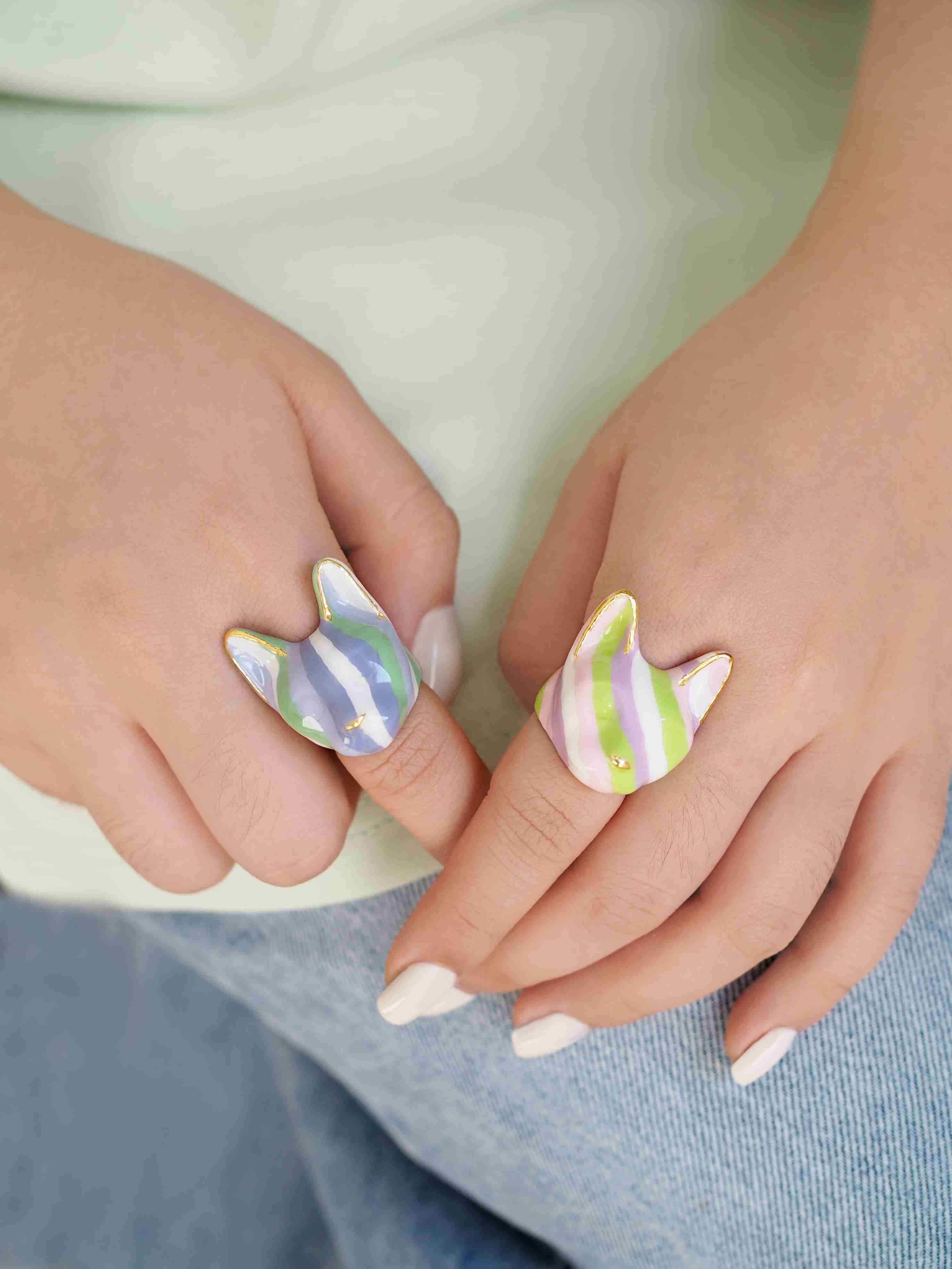 Two hands are shown wearing playful cat-shaped rings with soft purple, green, and white stripes, while the other features green, pink, purple, and white stripes.