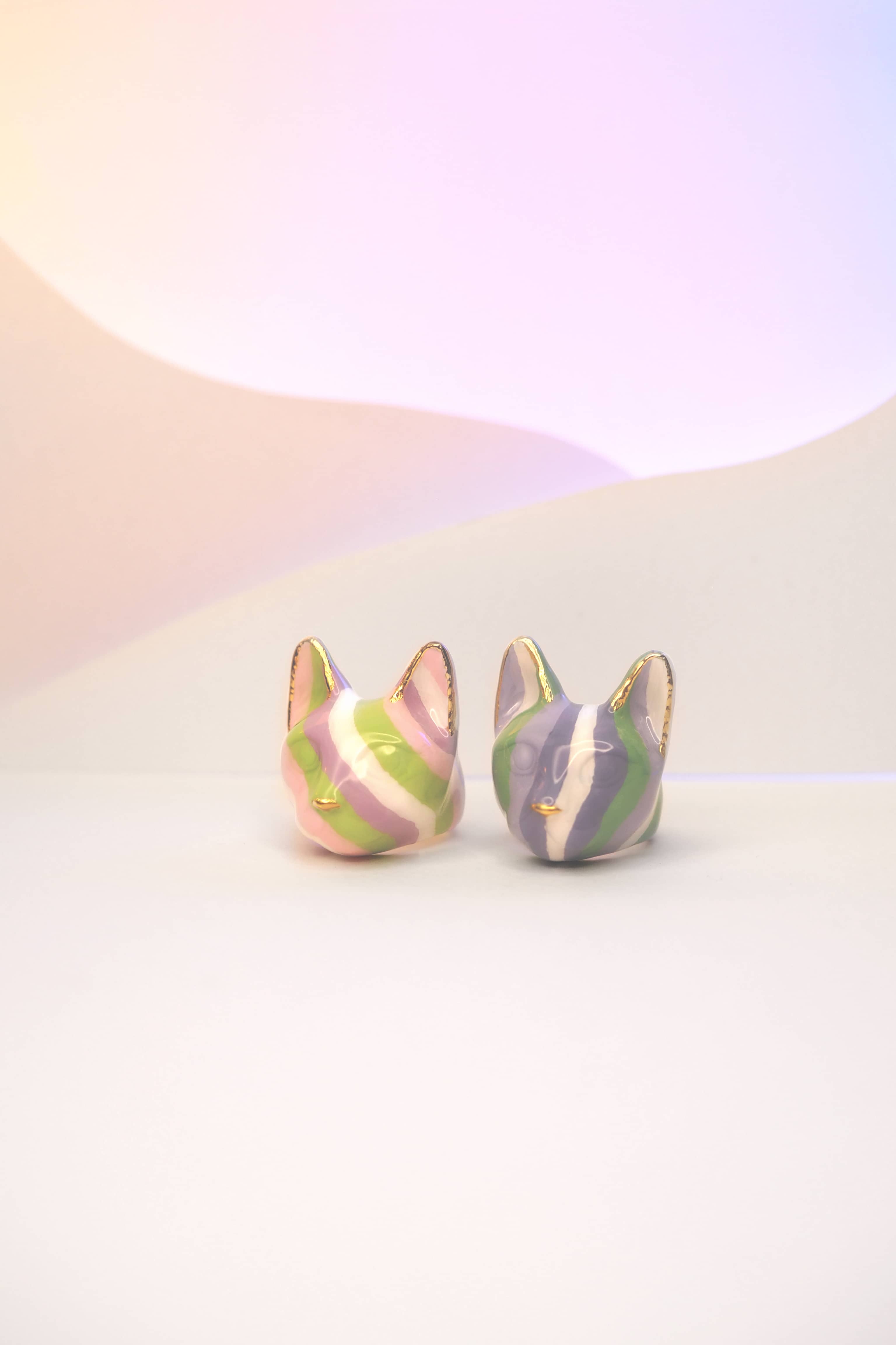 Two cat rings features alternating stripes of purple, green, and white, while the other with lilac, light green and pink set against a soft pastel background.
