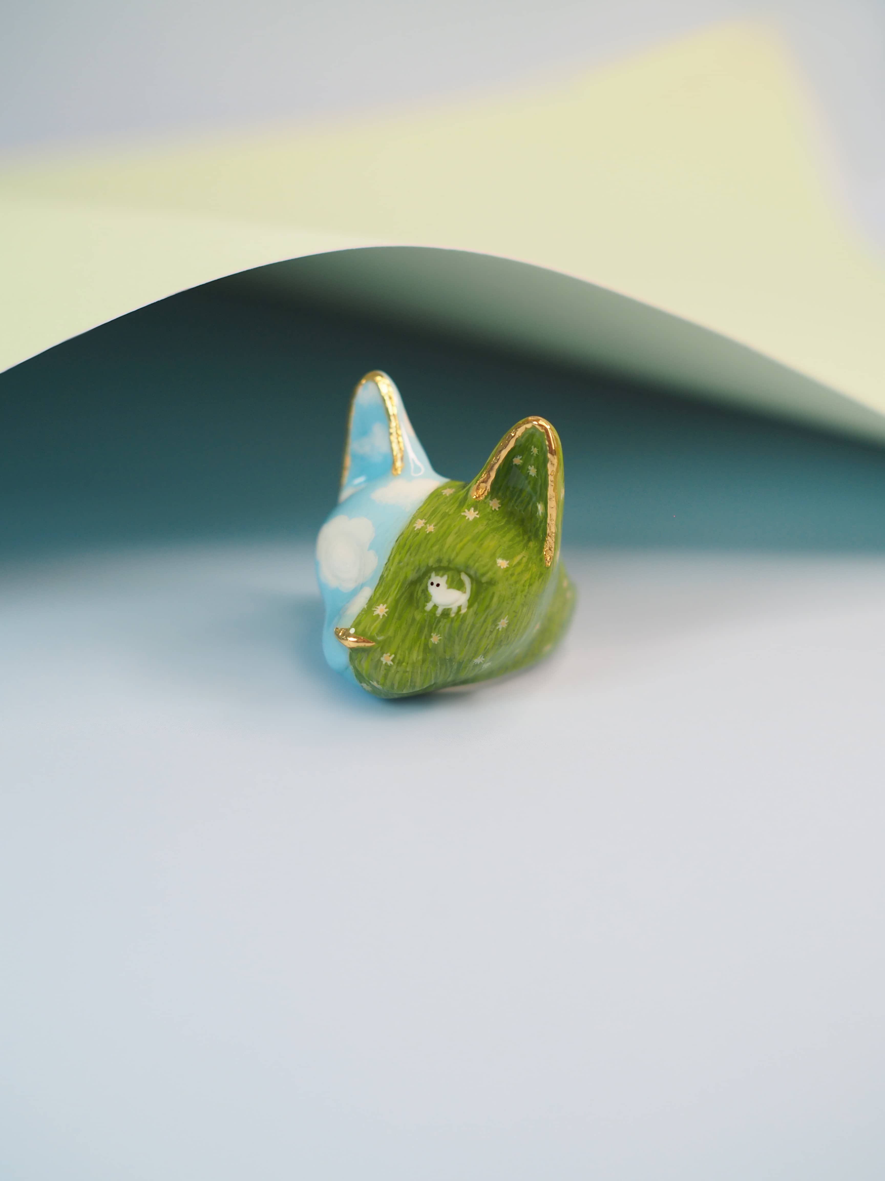 Artistic hand-painted cat ring with clouds and a grassy background.