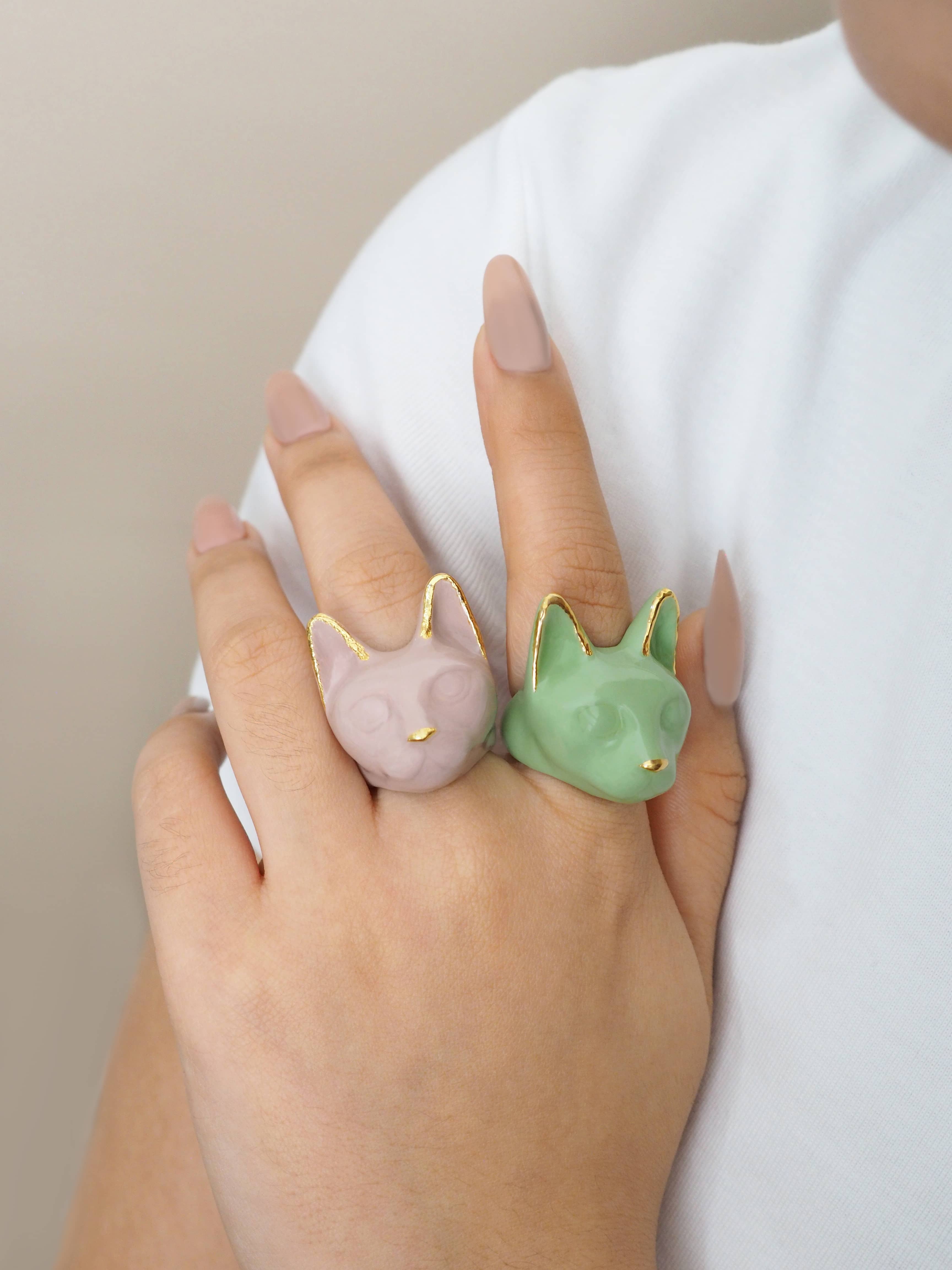 A hand wearing two cat rings: dusty pink and green with casual outfit.