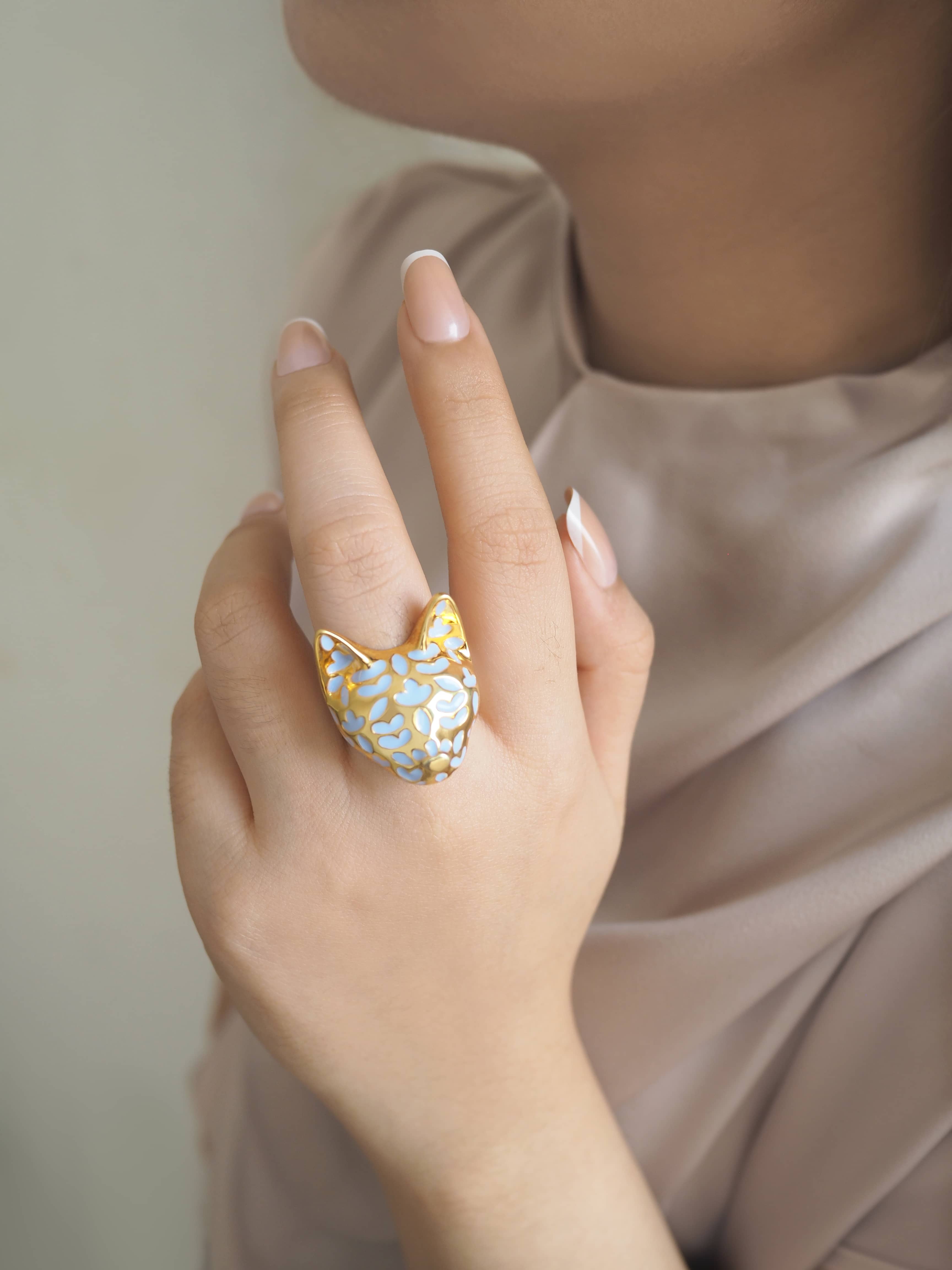 A close-up of a hand wearing a cat-shaped ring in purple and gold, inspired by lavender.