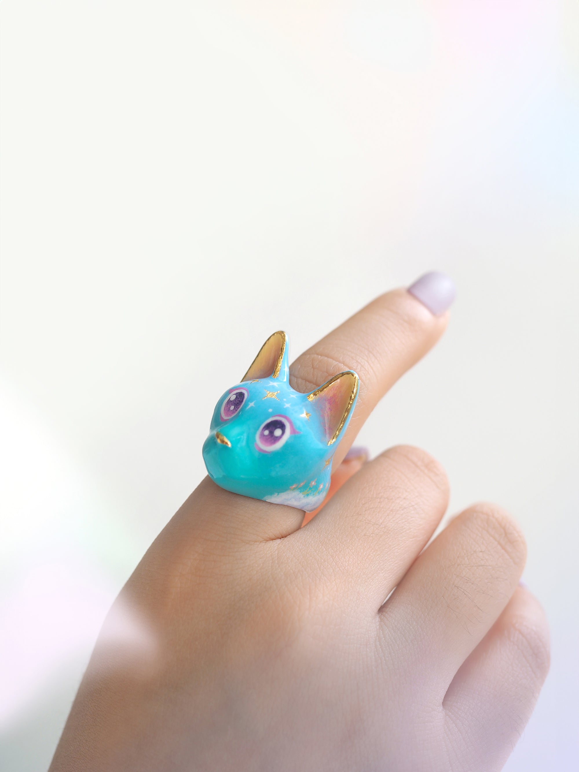 A close-up of a cat ring with in a soft blue color featuring sparkling purple eyes and star-like patterns on its forehead worn on a finger.
