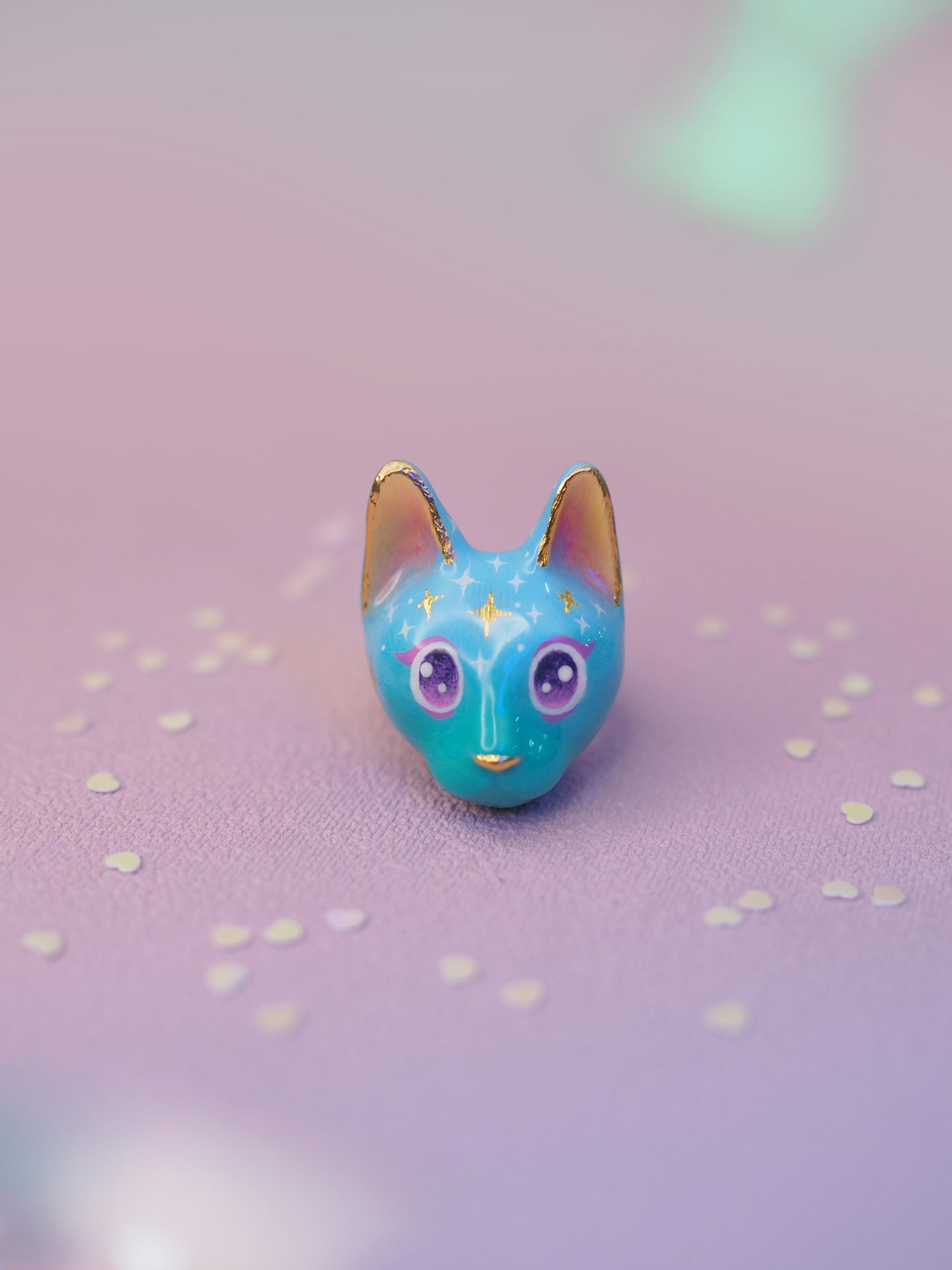 A whimsical cat-shaped ring featuring a blue face, yellow ears, sparkling purple eyes, and starry patterns placed on a light pink background.