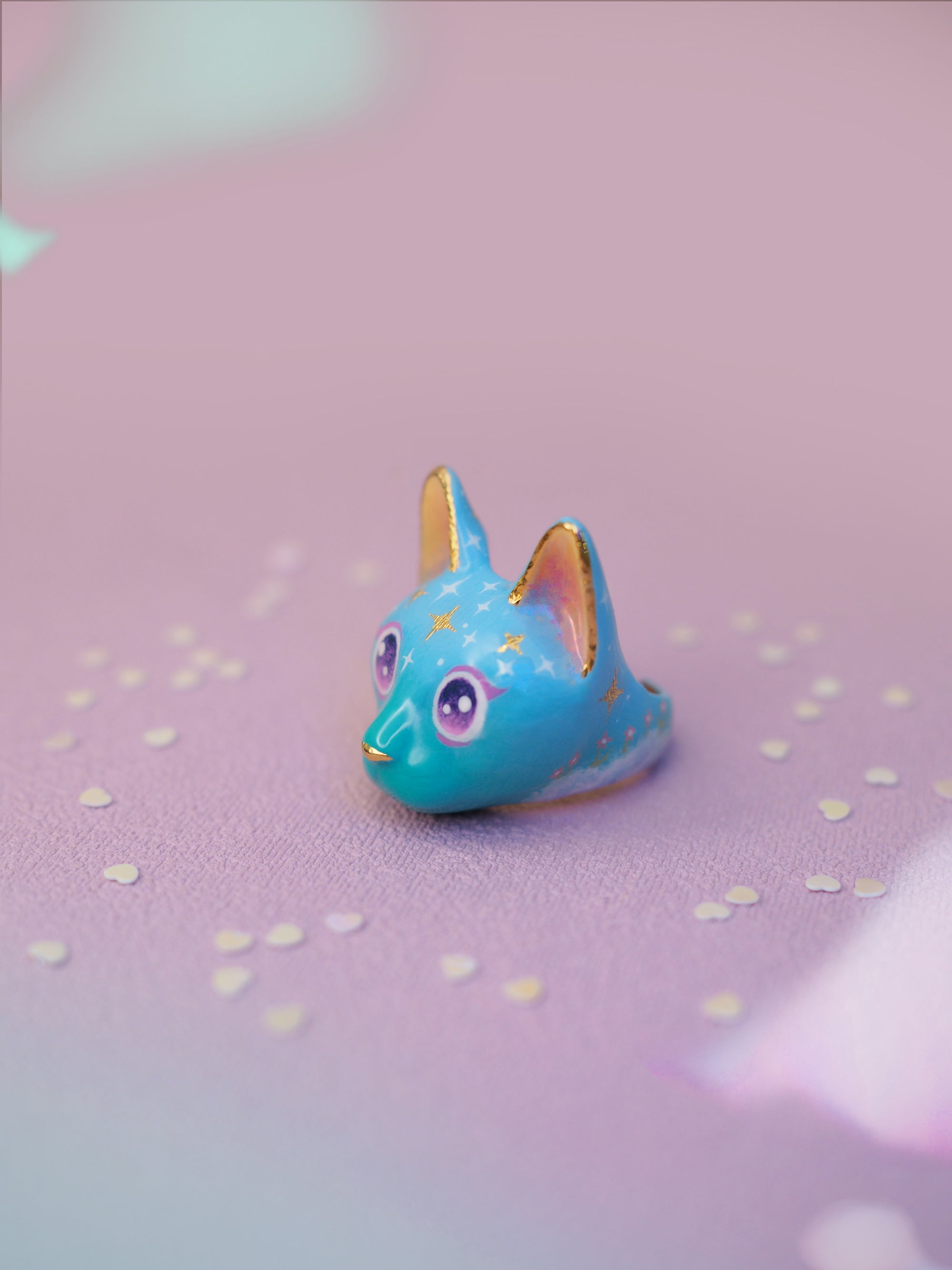Handcrafted cat ring featuring a blue face, yellow ears, sparkling purple eyes, and starry patterns placed on a light pink background.