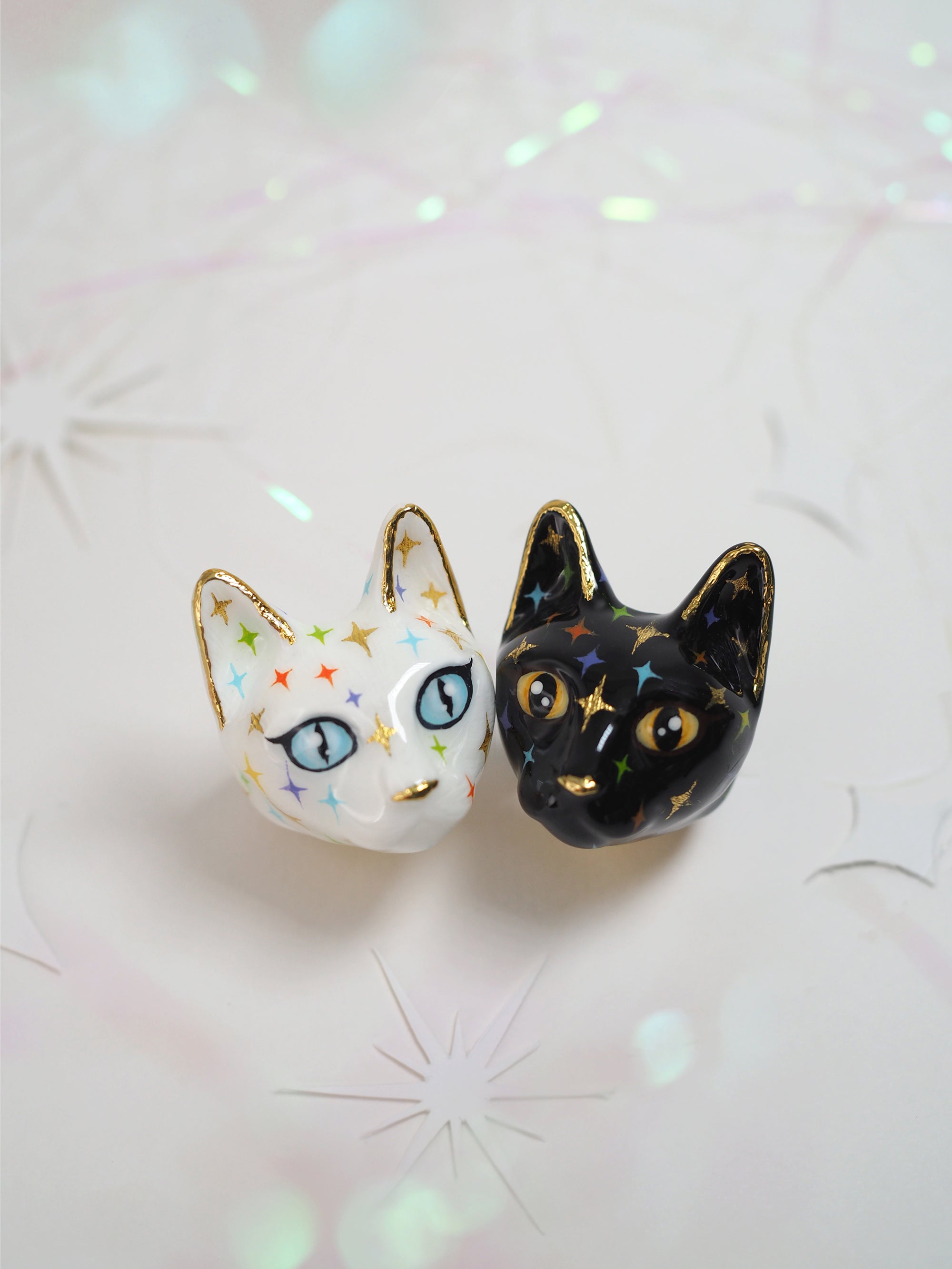Two whimsical cat face rings, one white with colorful twinkles and the other black with colorful twinkles.