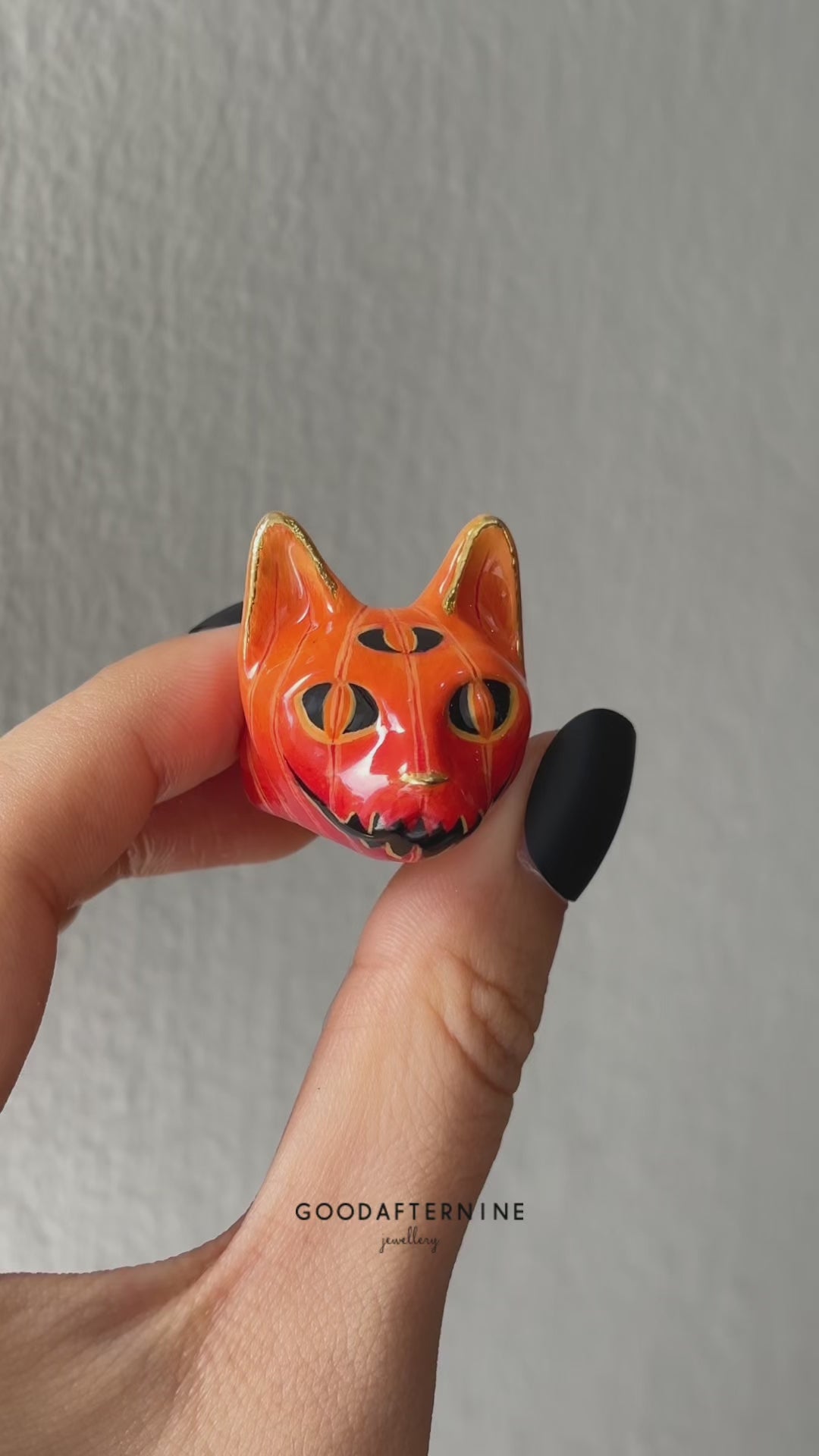 A close-up of a hand with black nail polish, adorned with two Halloween-themed cat rings a black cat with a skull motif and a pumpkin.