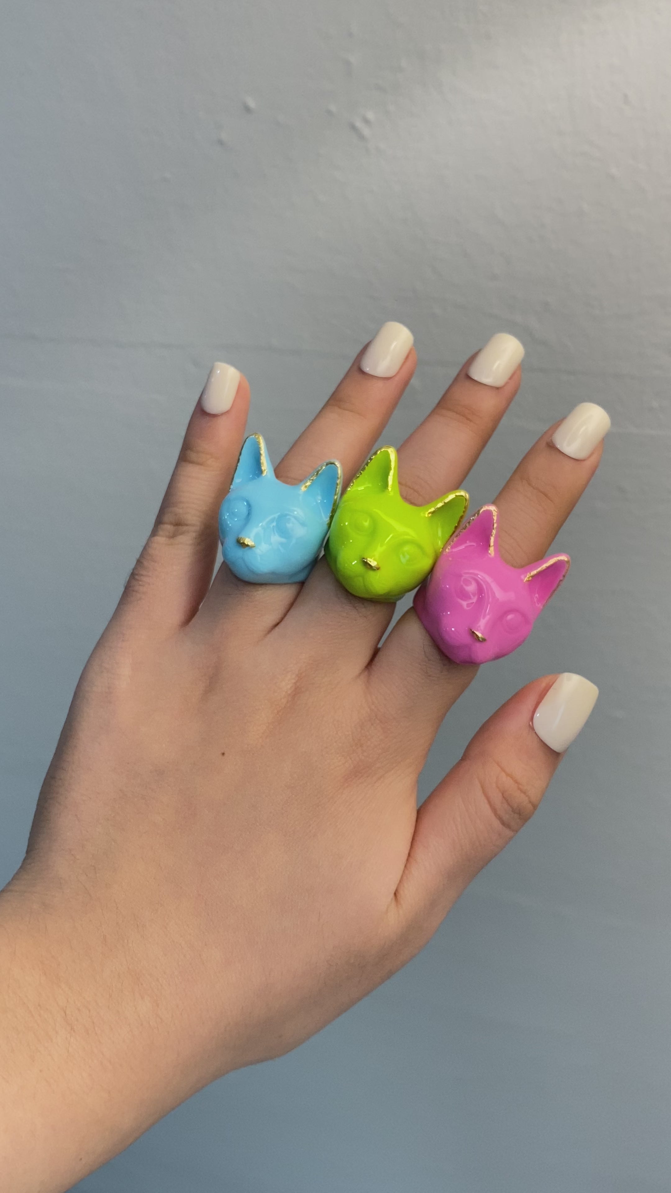 A hand wearing three colorful cat rings: pink, green, and blue.