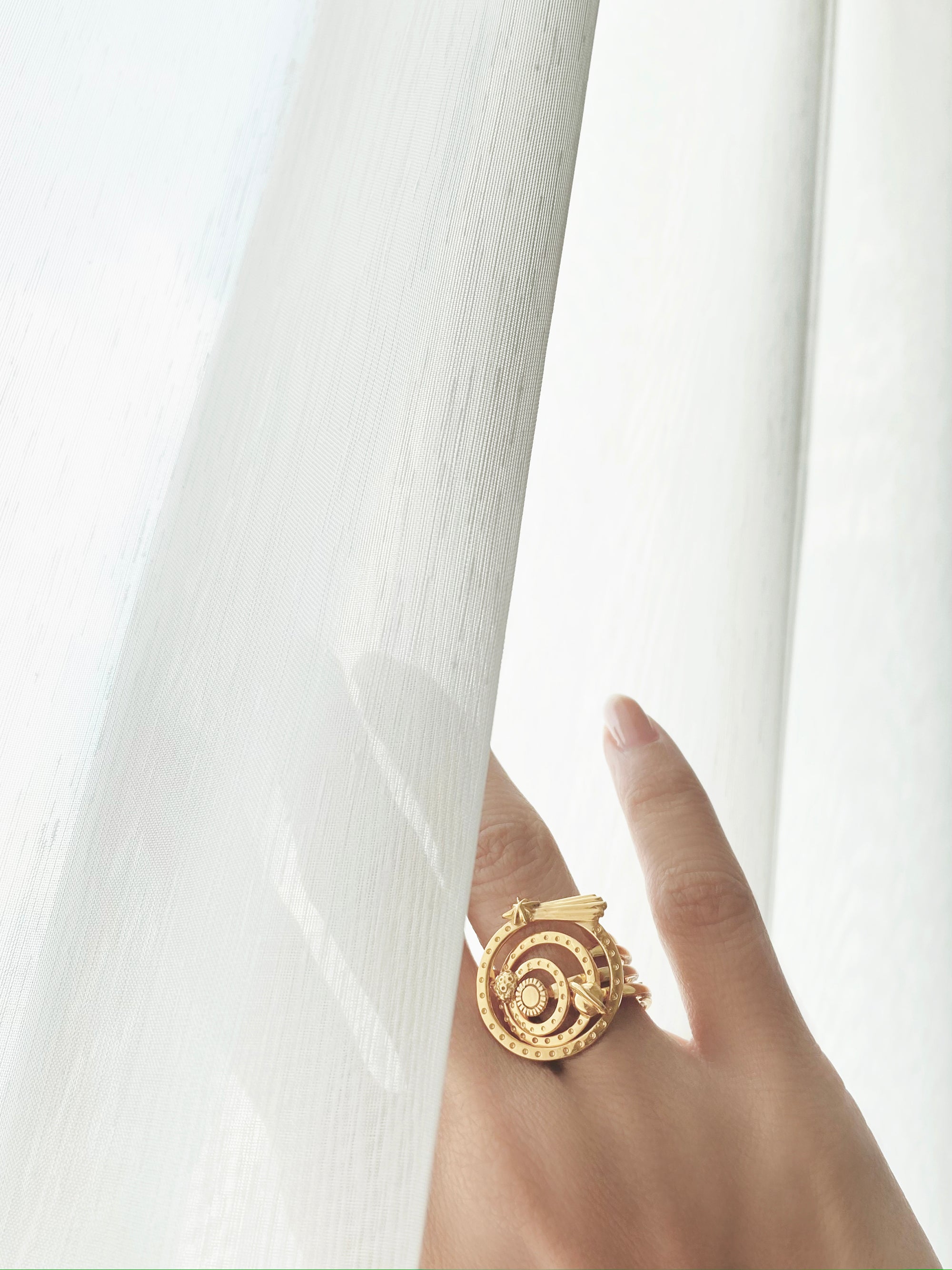Close-up of a hand adorned with solar system-inspired gold ring set hide behind the white curtain.
