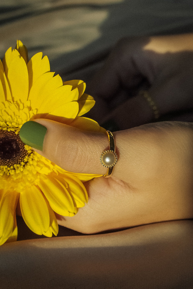 Gold ring with pearl inspired by the moon, worn on a finger next to a yellow flower.