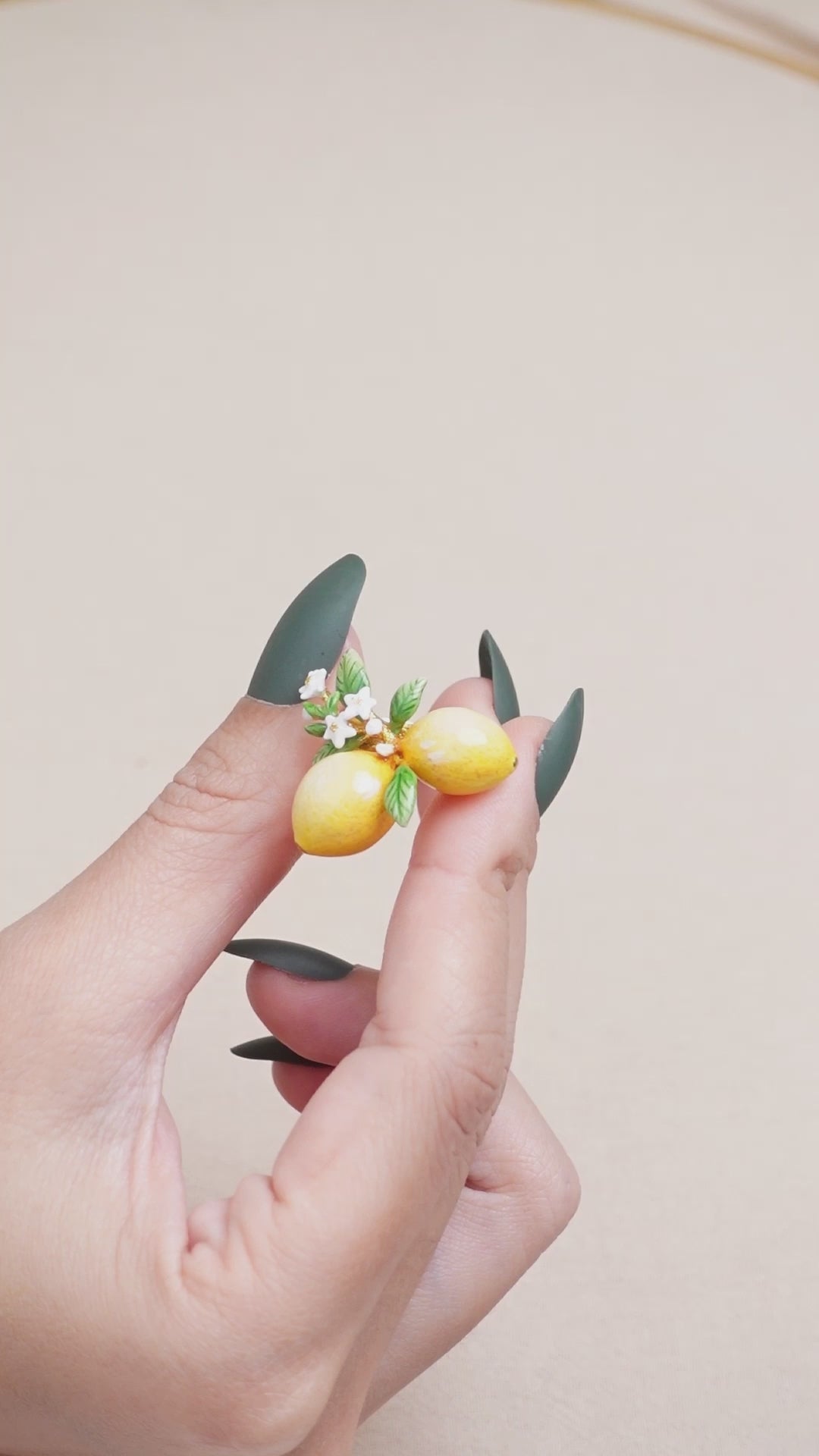 A hand wearing a handmade ring featuring two yellow lemons, green leaves, and small white flowers, ideal for summer fashion.