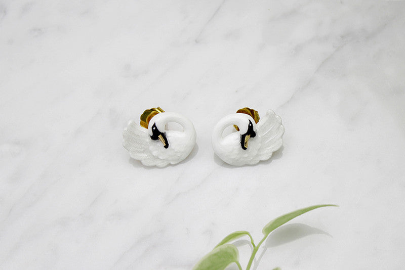 Pair of white swan-shaped earrings with black accents placed on a marble background with green leaves.