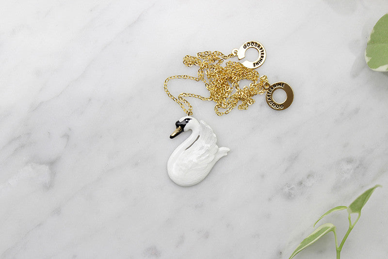 Beautiful swan pendant with gold chain necklace placed on a marble background with green leaves.