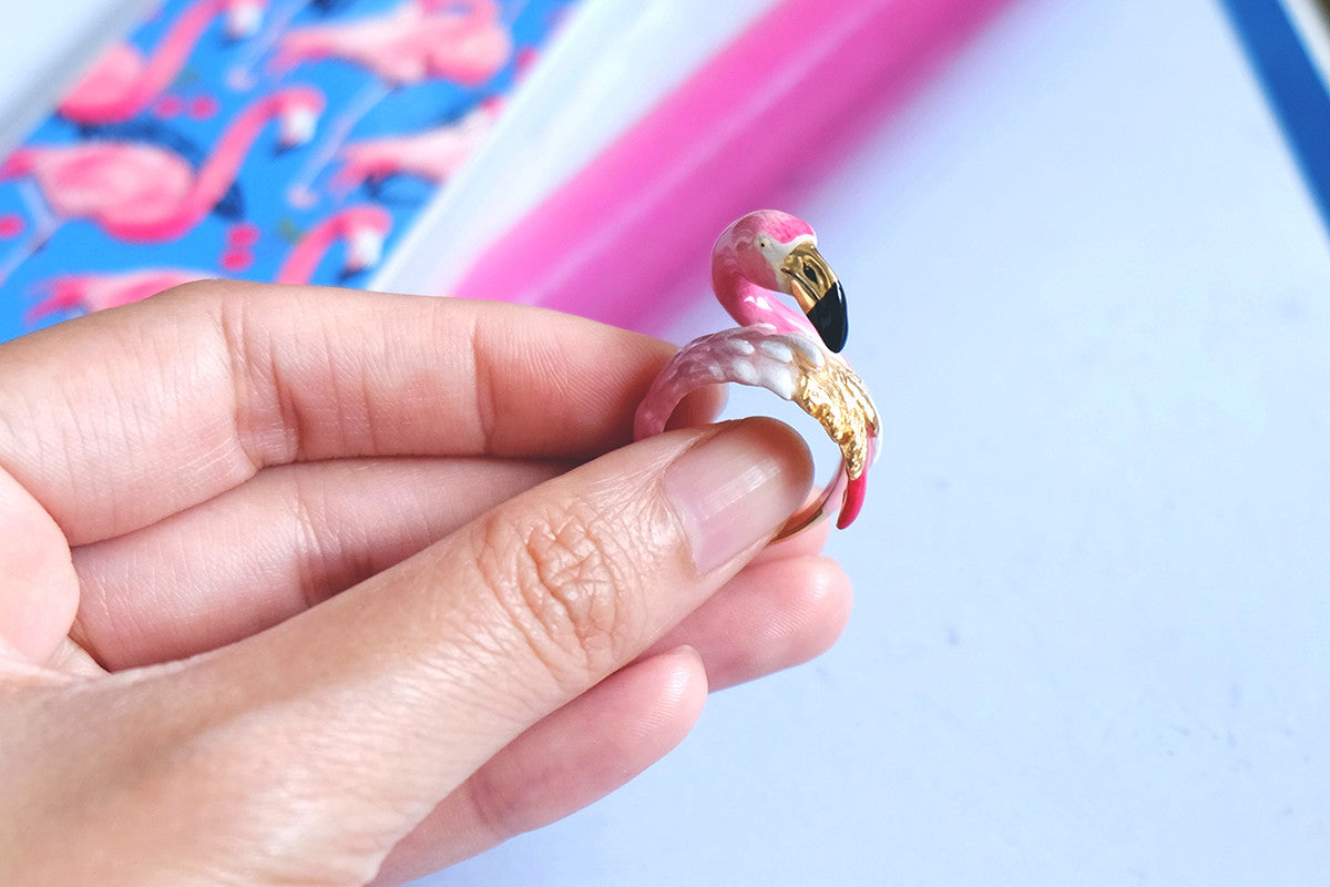 A hand holding a beautiful pink flamingo ring with a colorful background.