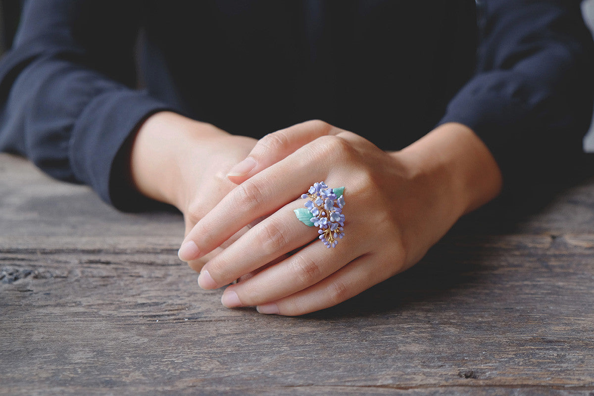 A hand adorned with a purple floral ring featuring lilac design rests on a wooden table.