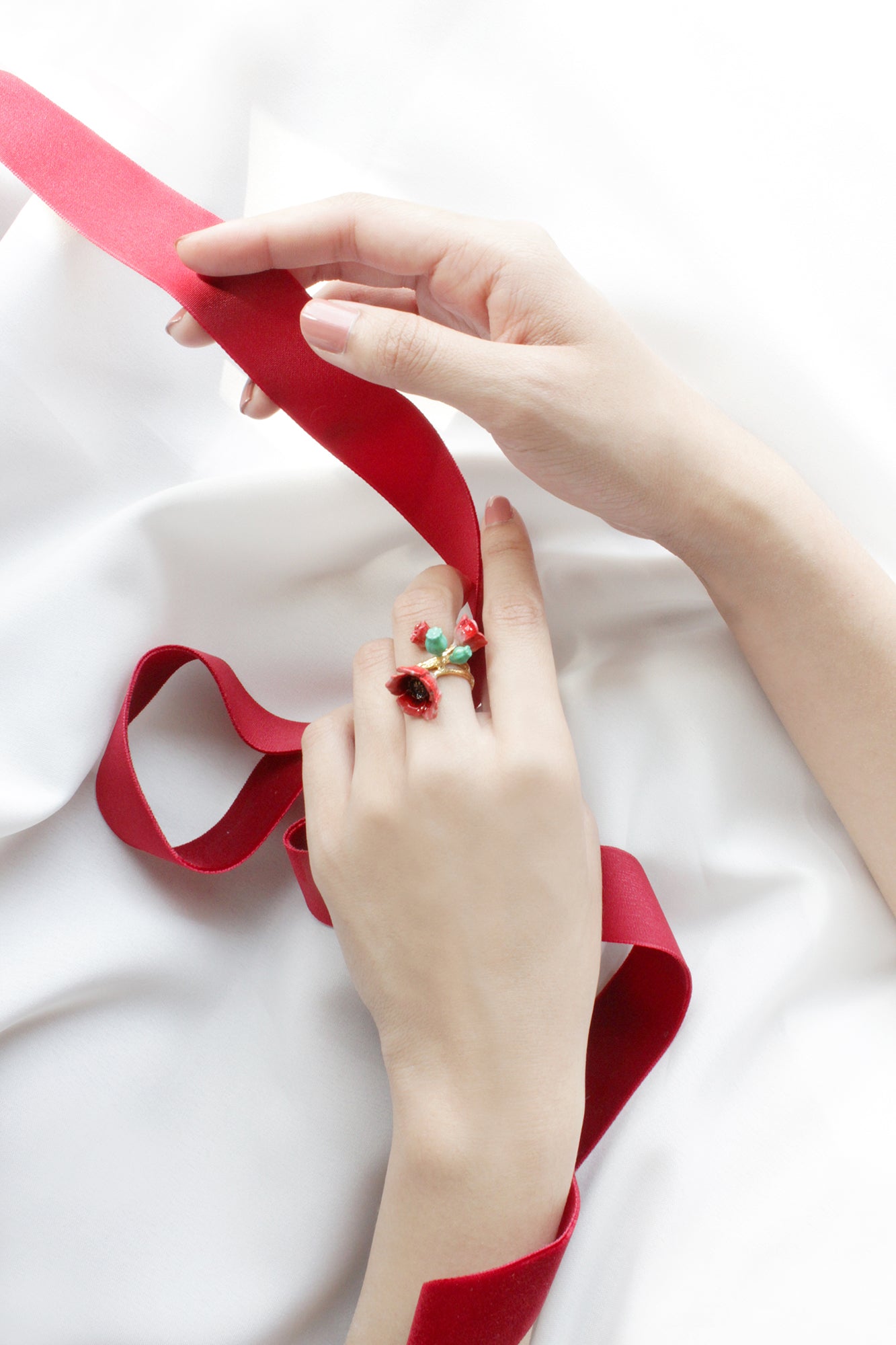A hand adorned with floral ring featuring red poppy design holding a red ribbon on a white fabric.
