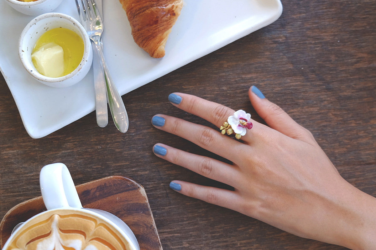A hand wearing a flower ring, featuring a white phalaen design, rests on a wooden table beside breakfast items.