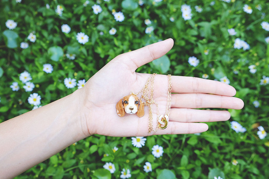 Close-up of a beagle pendant on a gold chain in a hand surrounded by flowers.