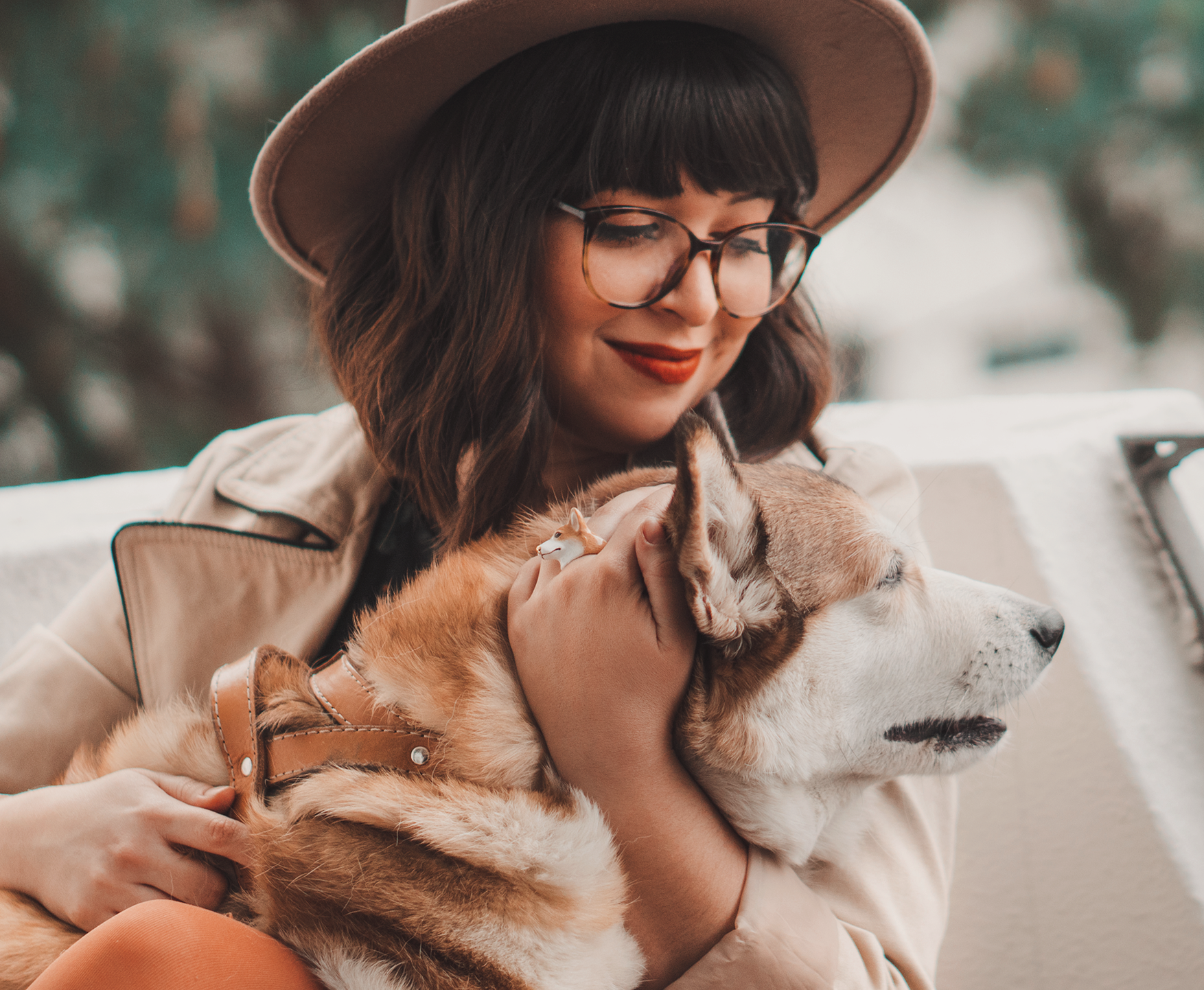 A woman with glasses, a hat, and a dog ring hugs her dog, enjoying a cozy moment together outdoors.